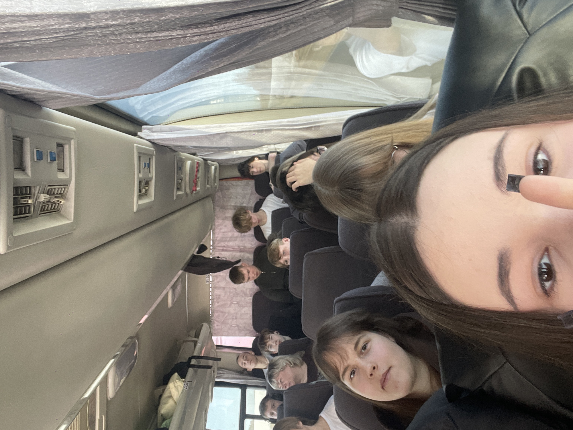 The image depicts the interior of a passenger bus, reportedly in transit within the city of Meshcheryaki, Russia. The perspective is a low-angle selfie, capturing approximately fifteen to twenty individuals, primarily young adults and teenagers, seated in rows.
In the immediate foreground, the upper portion of an individual's face is visible, including the eyes, forehead, and dark brown hair. A finger is shown adjusting a small, dark, rectangular object on the right eyebrow. Directly behind this individual, several passengers are seated. To the left, a young woman with brown hair, wearing a dark top, looks directly towards the camera. Behind her, an older woman with blonde hair and a dark top is partially visible. Further back, a young man in a dark hooded garment is seated prominently. Other young men and women are visible in various seats, some looking forward, others towards the camera, and some down. One passenger in the mid-background has visible earbuds.
The bus interior features dark grey fabric seats, light grey overhead compartments containing visible luggage items such as a white pillow-like object and a dark bag. The ceiling displays light fixtures, air vents, and passenger service units. The right side of the bus includes windows partially covered by patterned, light-colored curtains. The blurred view through the window indicates an outdoor environment consistent with open land or fields, suggesting the vehicle is in motion. No specific interactions between passengers are evident.
