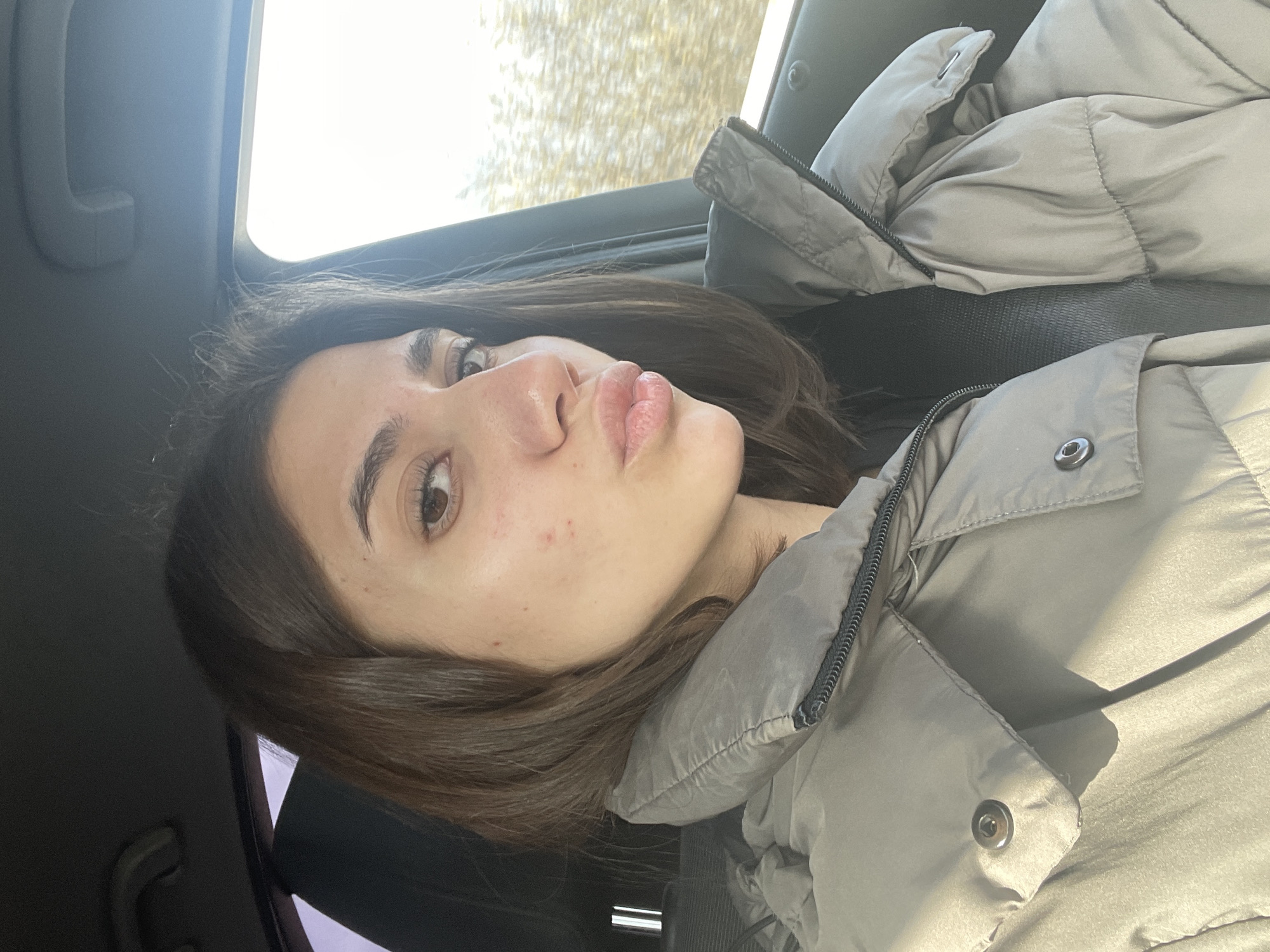 A young woman is in the passenger seat of a car, wearing a puffy, light gray jacket and a seatbelt. She has long, brown hair and brown eyes. Her lips are pursed as if she is posing for a selfie. The background shows a blurry, light-colored scene outside the car window, possibly trees or buildings on the outskirts of a city. The interior of the car is a dark color. The location is not apparent from the image.