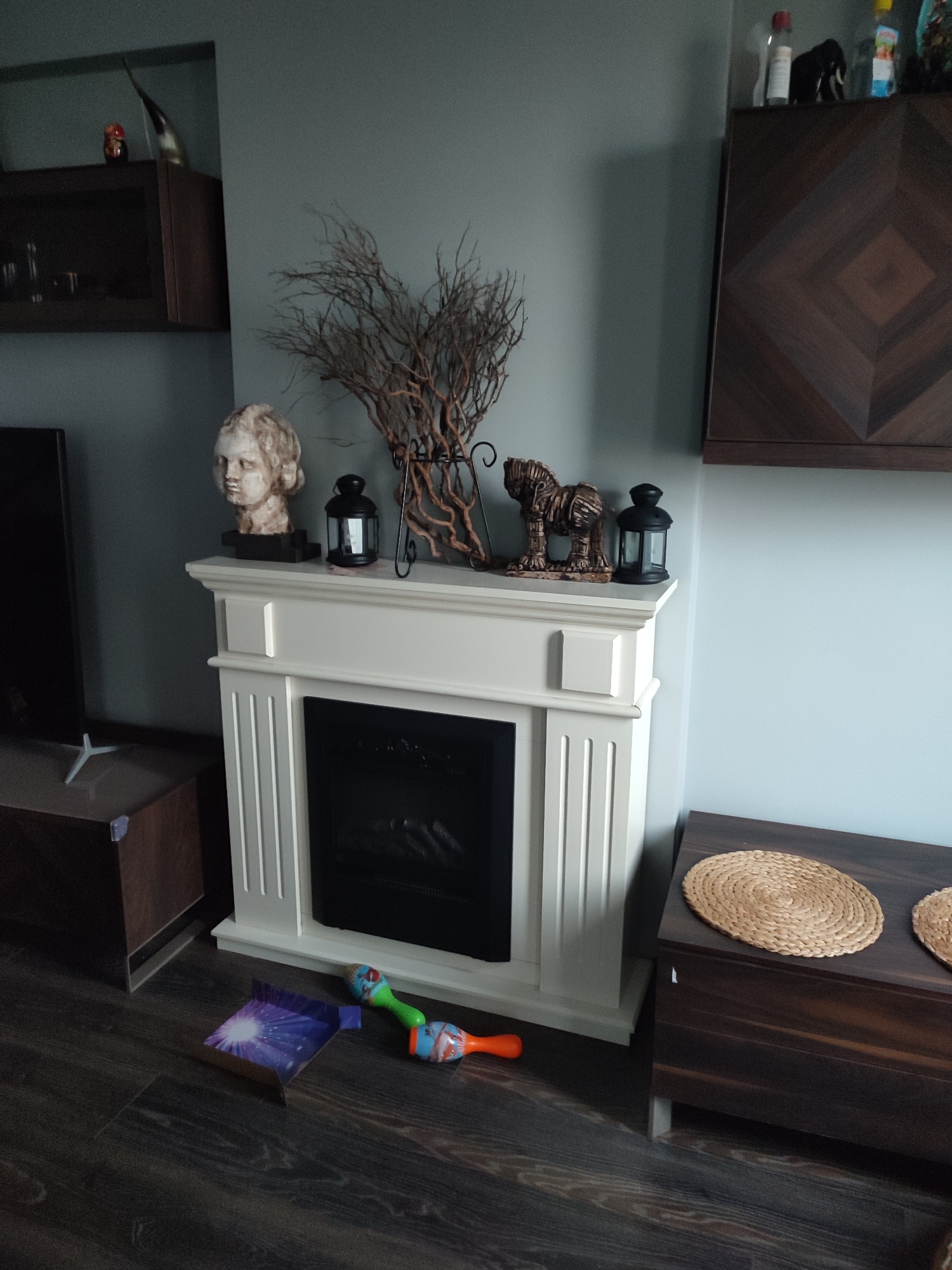 The image shows an indoor scene, likely a living room, featuring a white electric fireplace. On top of the fireplace are decorative items including a bust of a person, a branch arrangement, a statue of an animal, and two black lanterns. To the left of the fireplace is a dark-colored cabinet, and to the right is a wooden storage bench with two woven placemats on top. On the floor in front of the fireplace, there are children's toys, including a colorful maraca and a piece of cardboard with a picture of fireworks. The walls are painted a light grey-blue. The flooring is dark wood. There are no people visible in the image. The image does not contain any information that would indicate its location in Kartal, Turkey.