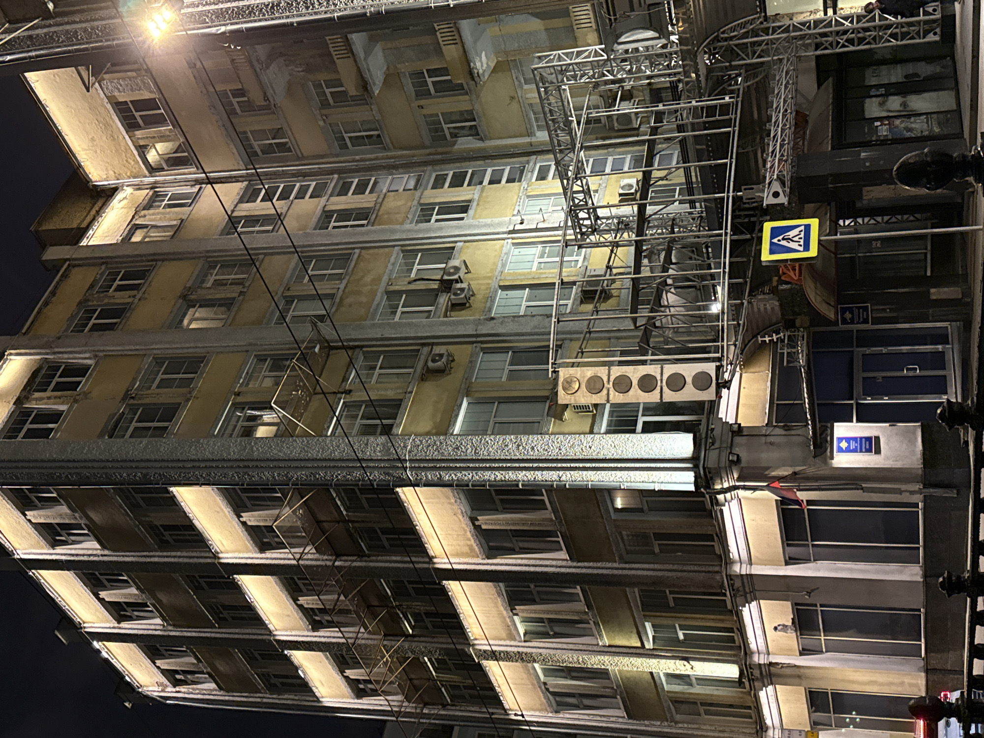 The image depicts an eight-story building in Moscow, Russia, as evidenced by the street sign with the pedestrian crossing symbol. The building appears to be undergoing renovation, indicated by the scaffolding covering the façade. The building's exterior is primarily yellow and gray with numerous windows. A streetlight illuminates the scene. Air conditioning units are visible on the building's exterior. A blue plaque is affixed to the building's right side. The background suggests a nighttime setting.