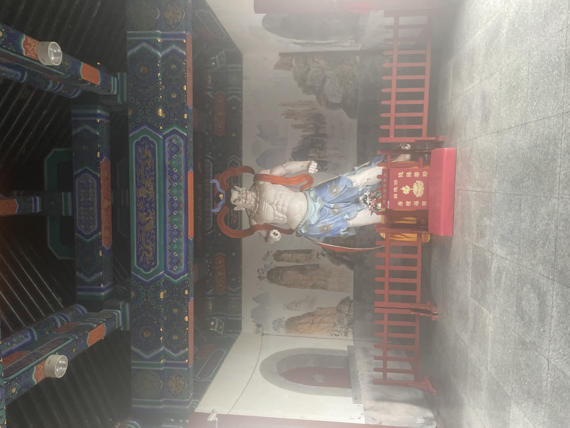 The image shows a statue of a muscular figure with red headgear, possibly a deity, positioned in what appears to be a temple or shrine within the Yongding District, China. The statue is situated behind a red wooden fence and rests on a red stand with Chinese characters inscribed on it. The backdrop features a mural depicting a landscape with mountains. To the left of the statue, there's a section of ornately decorated architecture with colorful beams and columns. The environment appears hazy or foggy, obscuring some details of the scene.