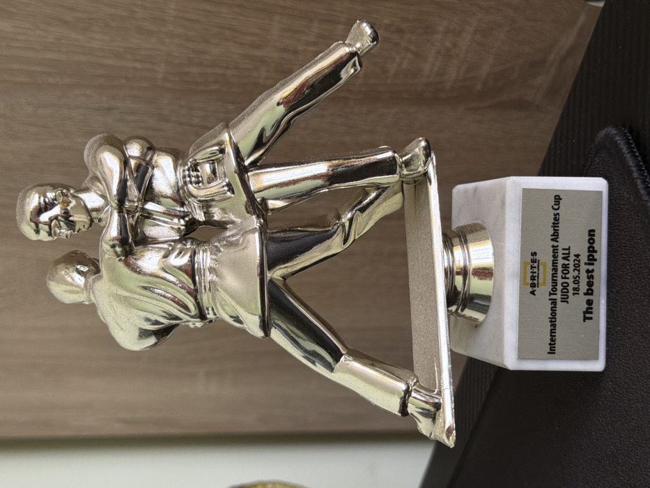 A silver-colored trophy, depicting two stylized male judoka grappling in a dynamic throw or technique, is prominently displayed. The figures wear white judogi and dark belts, with their limbs and torsos forming an open, active composition. They stand on a horizontal silver-colored bar, which is supported by a central silver column resting on a rectangular white marble base. A rectangular metallic plaque is affixed to the front of the base, bearing the "A-BRITES" logo in the upper left corner. The inscription on the plaque reads: "International Tournament Abrites Cup", "JUDO FOR ALL", the date "18.05.2024", and "The best ippon". The trophy is situated on a dark, textured surface, possibly a desk, against a background of a light brown, vertically-grained wooden panel on the right, and a lighter, plain surface on the left. Based on the "Abrites Cup" inscription and the known location of the company Abrites, the tournament and trophy are associated with the city of Sofia, Bulgaria.