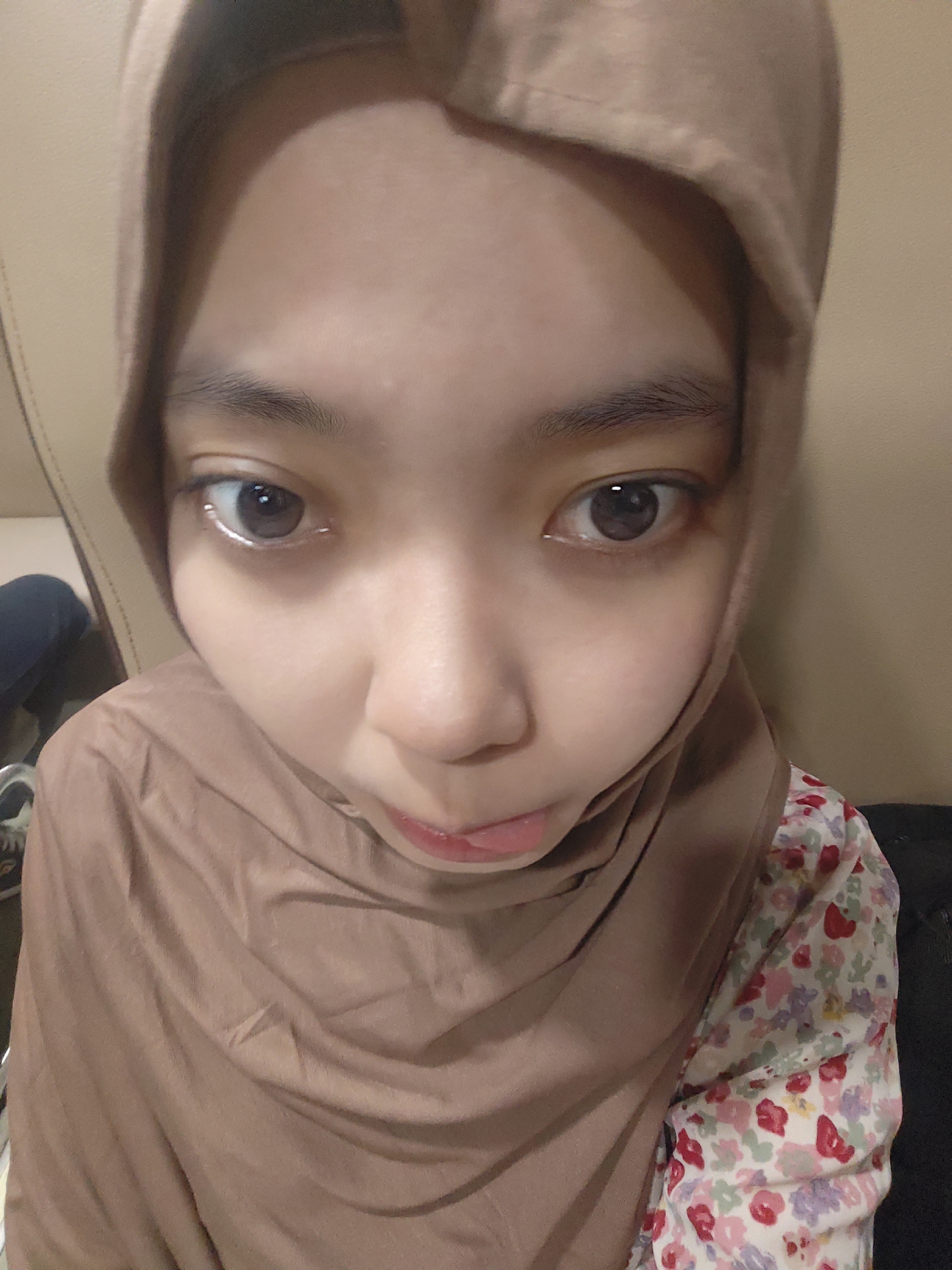 The image shows a close-up, slightly distorted, selfie of a young person wearing a brown hijab and a floral patterned shirt. The person's face is the primary focus, with their eyes wide open and looking directly at the camera. The background appears to be indoors, with a textured, possibly leather, surface behind the person, suggesting a couch or chair. There is also a hint of another person or object in the background to the left, which is out of focus. The lighting is somewhat dim, creating shadows on the person's face and the hijab. No specific landmarks or details that would indicate the location in Plandaan, Indonesia, are visible.