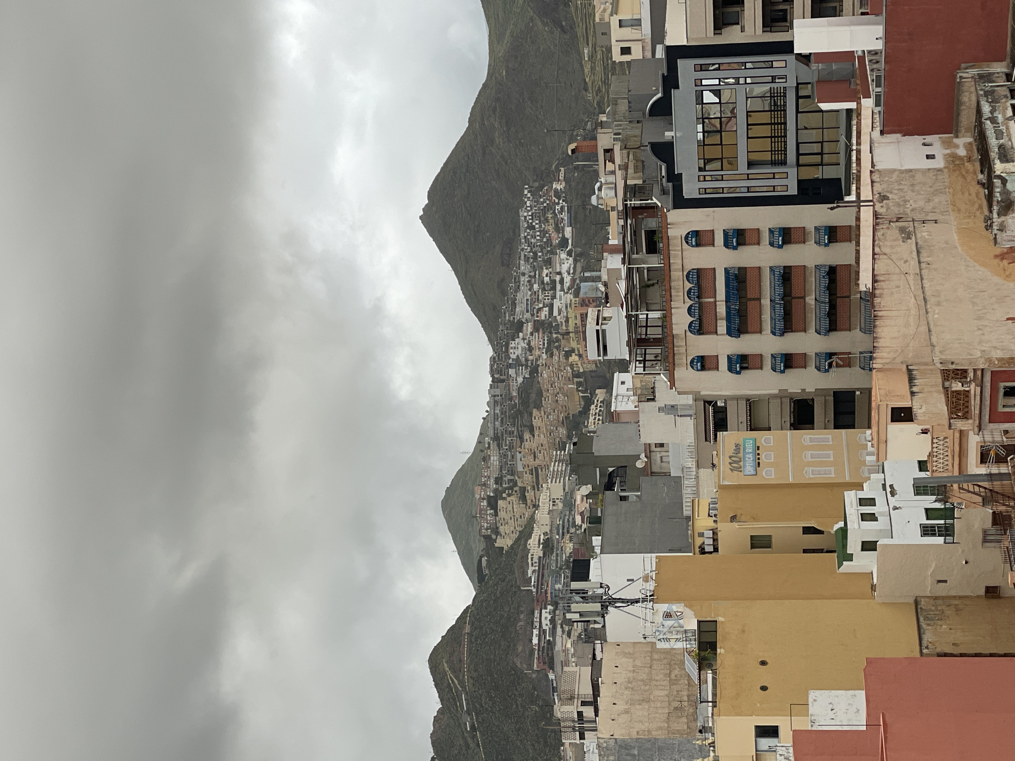 The image presents a panoramic view of a densely populated city nestled against a large, verdant mountain under an overcast sky. The scene is set in Santa Cruz de Tenerife, Spain, evident from the distinctive mountainous terrain and Mediterranean-style architecture.
The foreground and midground are dominated by numerous buildings of various heights and designs, mostly multi-story structures with flat roofs. Colors range from traditional whites, creams, and earthy tones like terracotta to more modern grey and black facades. Many windows feature blue or brown shutters and balconies. Rooftops display an array of satellite dishes, air conditioning units, and what appear to be solar panels. Electrical wires and poles are also visible throughout the urban sprawl. The city extends upwards, covering the lower slopes of the steep, green mountain, which forms a dramatic backdrop.
The weather is cloudy, with heavy grey clouds dominating the sky, suggesting a cool, possibly rainy day. The light is diffused, indicating daytime without direct sunlight. No people or specific activities are visible; the scene depicts a static urban and natural landscape.
A notable detail, visible in one of the provided crops, is a sign on a yellow building that reads: "100 años OPTICA RIEU". This indicates a long-standing optical business. The overall impression is one of a vibrant, compact city integrated into a striking natural environment.