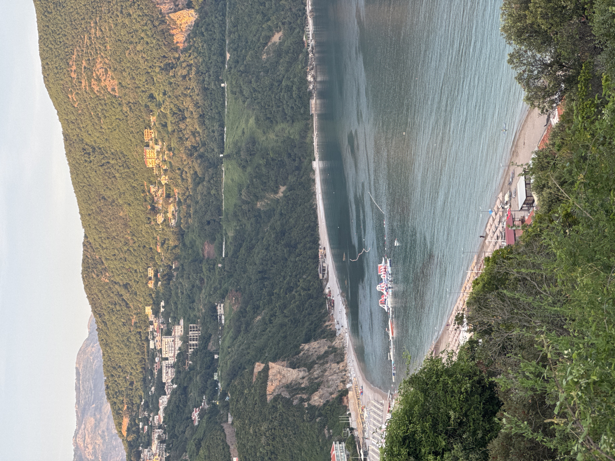 The image shows a view of Prijevor beach in Budva, Montenegro. The beach is a narrow strip of sand along the coast. A variety of leisure craft including boats and floats are in the sea. The shoreline is lined with sunbeds and umbrellas. Buildings of various styles and sizes are scattered along the edge of the beach and nestled into the wooded terrain that rises steeply. The slopes are densely covered with green trees and shrubs. In the distance, there are high mountains. The sea surface shows gentle ripples. The sky is overcast, creating a soft and diffused light.