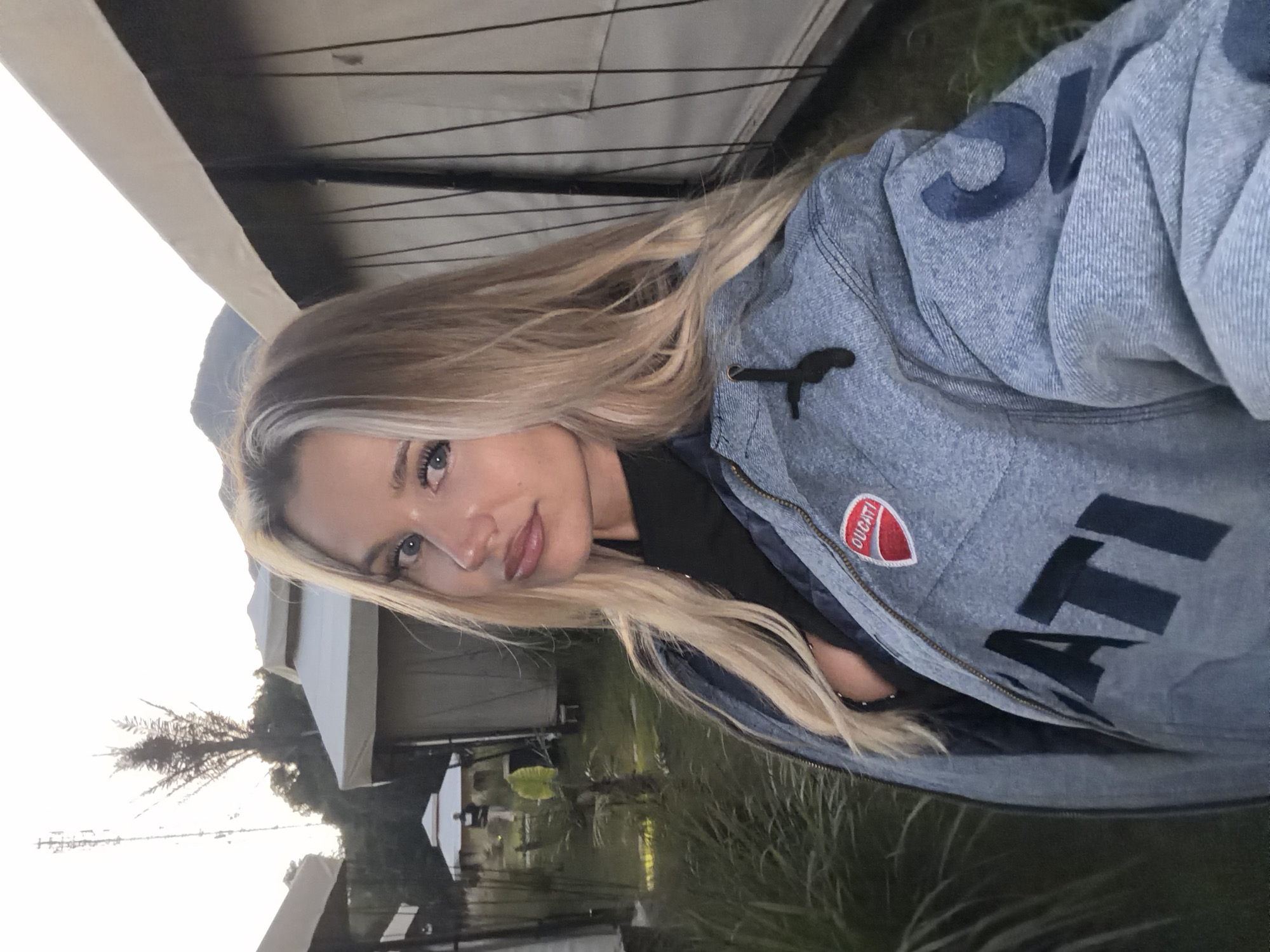 A blonde woman with blue eyes, wearing a black top and a denim jacket with the word "DUCATI" emblazoned on it, is taking a selfie outdoors. The jacket has a Ducati logo patch on the left chest. She is standing in what appears to be a glamping site with tents in the background. Lush green grass and palm trees are visible. In the distance, a telecommunications tower stands against a bright sky. The scene is set in Kintamani, Indonesia, characterized by its mountainous terrain and lush tropical vegetation.