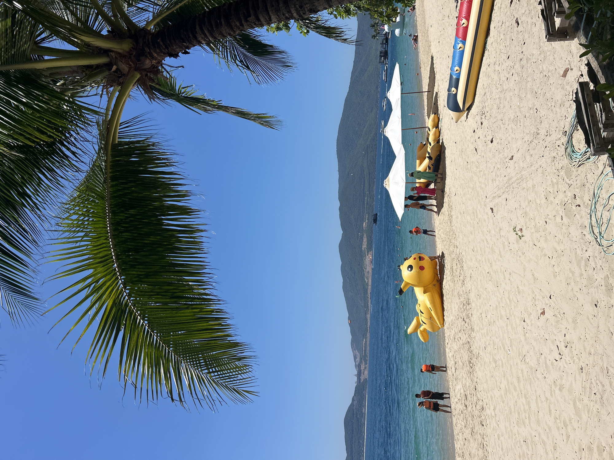 This image depicts a sunny day at the beach in Bac Nha Trang Ward, Vietnam. The foreground is dominated by a sandy beach with various objects scattered on it. A large, yellow inflatable Pikachu figure lies on its side near the water's edge. Several people, some wearing life vests, are gathered on the sand and near the water. Three white beach umbrellas are set up on the sand, providing shade. To the right, a banana boat, colored yellow, blue, and red, is resting on the sand. In the background, the turquoise ocean stretches towards a coastline lined with green, mountainous terrain. Several boats and a distant pier are visible on the water. Palm fronds from a large palm tree partially obscure the top right portion of the image, framing the clear blue sky.