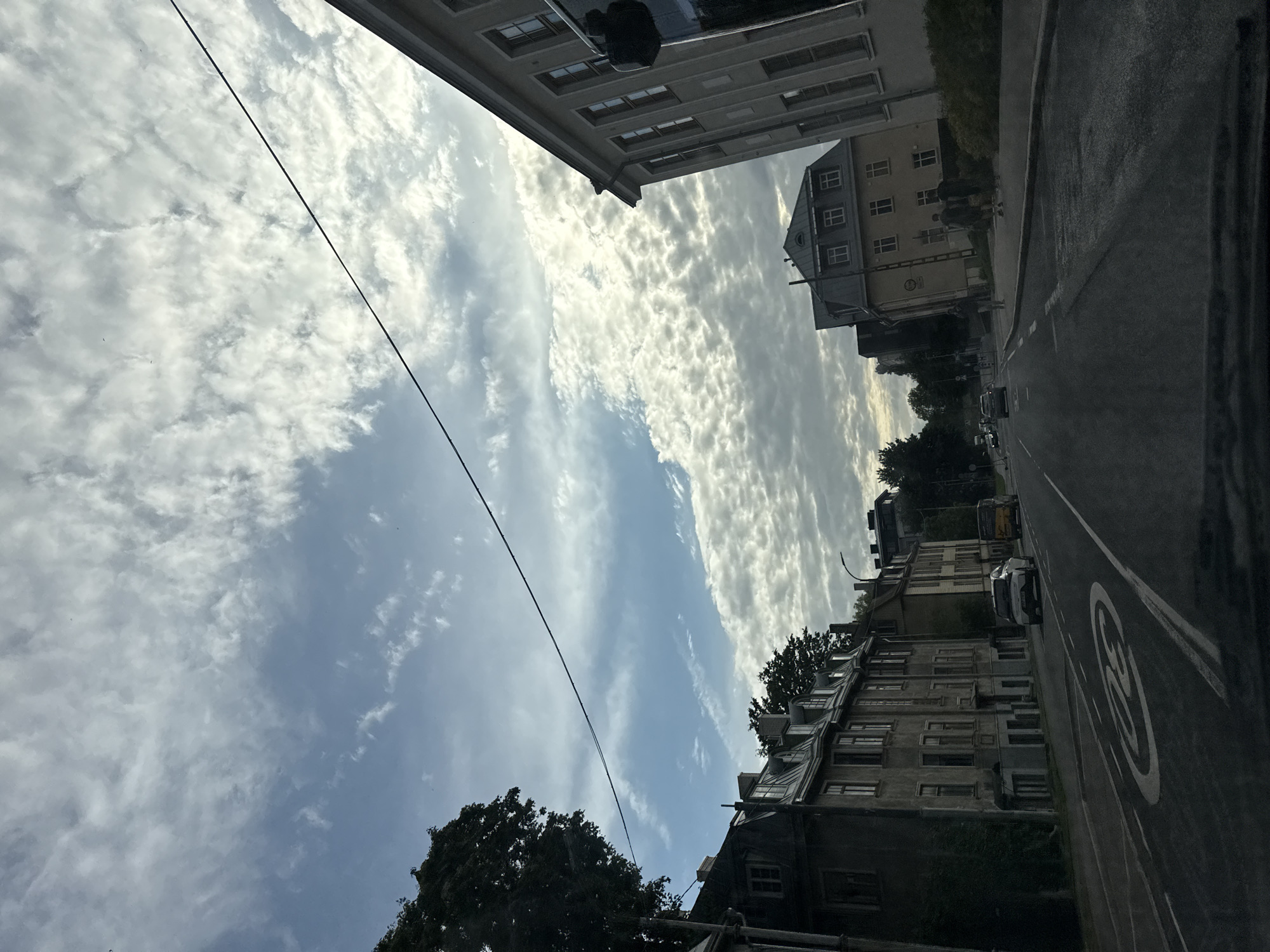 The image depicts a street scene in Tallinn, Estonia, likely on a cloudy day. The perspective is from inside a vehicle, looking forward and slightly upwards. In the foreground, the asphalt road surface is visible with a white circular speed limit sign indicating "30". On the left side of the road, there are several old, multi-story buildings with mansard roofs, some covered in ivy. A large tree with dark foliage is present on the far left. Across the street, on the right, is a lighter-colored, modern building with a series of windows. Further down the street, more buildings are visible, including a yellow building with a dark, steep roof. Several cars are on the road, including a white SUV and a yellow van. In the distance, the road curves and disappears behind buildings and trees. The sky is filled with a mix of blue sky and scattered, fluffy white clouds, creating a dynamic cloudscape. A thin black wire stretches diagonally across the upper portion of the image. There are no identifiable individuals prominently featured in the scene.
