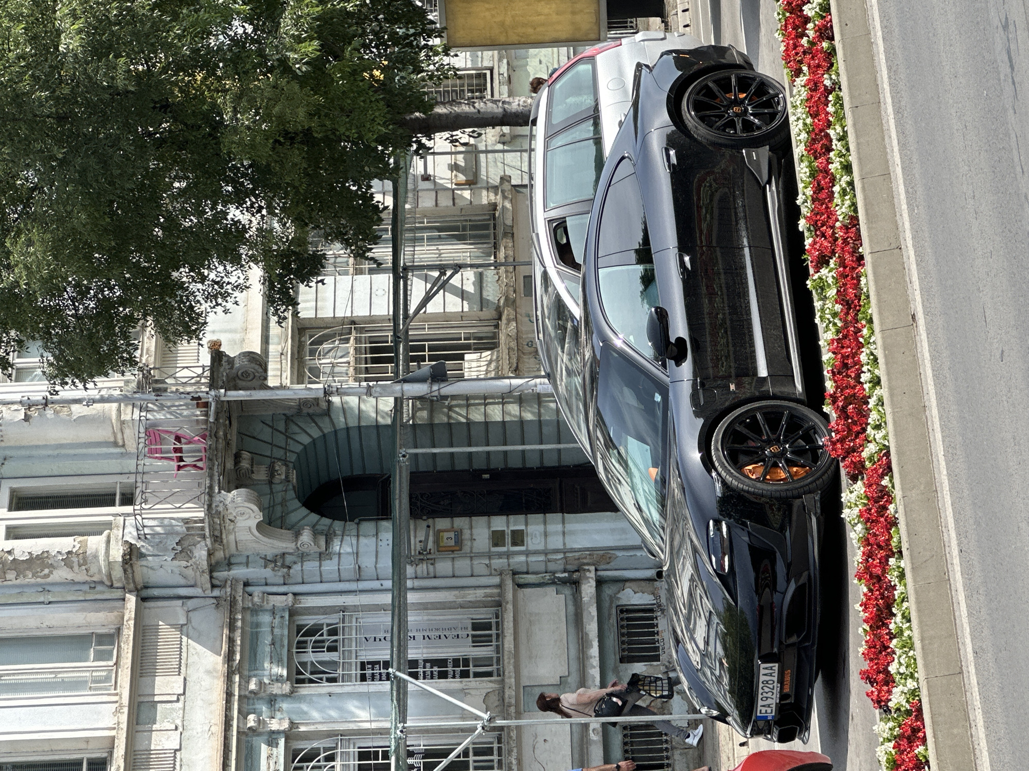 The image depicts a street scene in Varna, Bulgaria, featuring a black Brabus car parked alongside a curb decorated with alternating red and white flowers. The car's license plate reads "EA 9328 AA". Behind the car is a white vehicle and a large building with scaffolding. Several people are visible, including individuals walking near the scaffolding. The building's architecture is aged, with peeling paint and ornate details. A tree's foliage partly obscures the building's upper section. The text "АСТОЛИТА SC", "BRABUS", "DECOPA", "ПАЗАРНИ", "АЛИЗИ", "РИДИЧЕСКА", "MOU", "ОДАЖБА", "ОЧА", "СЕДЕМ К", and "недвижими и" are visible on signage near the building.