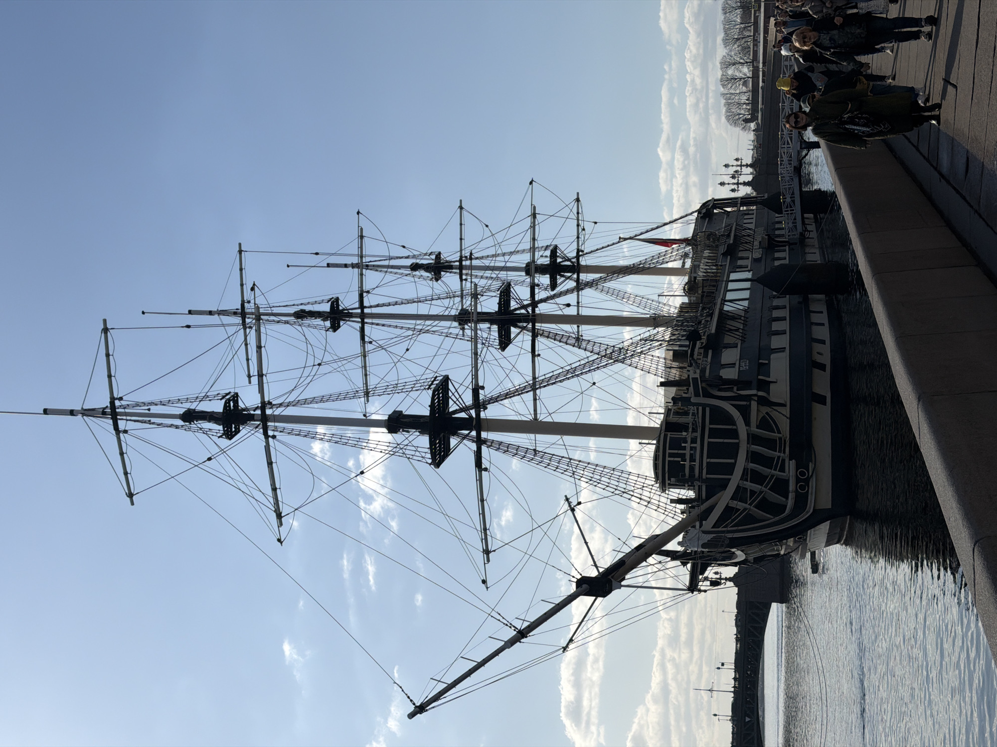A three-masted sailing ship, painted in dark and light tones, is permanently moored on the Neva River in Saint Petersburg, Russia, adjacent to a gray stone embankment. This vessel is identified as the "Flying Dutchman" restaurant ship. Its hull is predominantly dark, featuring lighter decorative elements on the stern and simulated gunports. Extensive rigging and spars define its silhouette against a clear blue sky with scattered cumulus clouds. A red ensign with a white cross is visible flying from the stern of the vessel.
In the lower right foreground, a group of approximately ten pedestrians walks from right to left along the stone embankment. They are dressed in casual coats and jackets, with their faces mostly indistinct or obscured. To the immediate left of the ship's bow, the Birzhevoy Bridge, a green metal arch structure, spans the river. Distant deciduous trees, currently bare, are visible along the far riverbank. The scene is illuminated by strong sunlight, casting visible shadows from the pedestrians and the embankment onto the walkway.