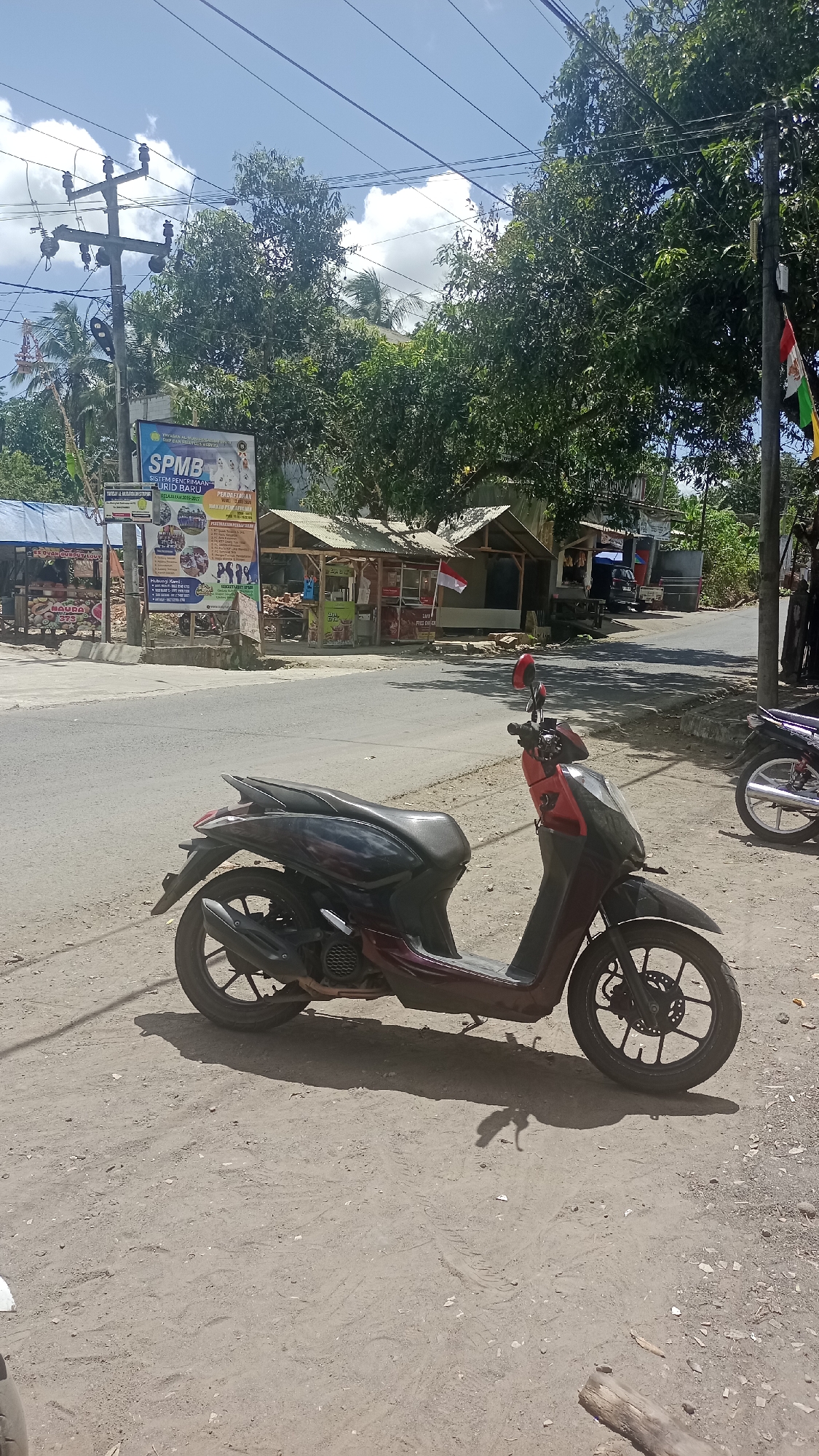 The image depicts a street scene in Cibeber, Indonesia, under a bright, cloudy sky. In the foreground, a dark red and black scooter is parked on a dirt shoulder next to an asphalt road. The scooter is angled towards the right of the frame. The background reveals several small roadside structures, likely food stalls, with awnings and open fronts. A large banner with text in Indonesian and images of people is prominently displayed on the left side, near a utility pole. Trees with lush green foliage line the area behind the stalls and along the road. A small Indonesian flag, red and white, is visible flying near one of the stalls. The road appears to be a two-lane street with some traffic. The overall impression is a typical day in a local Indonesian town, with the scooter and roadside commerce being central elements of the scene.