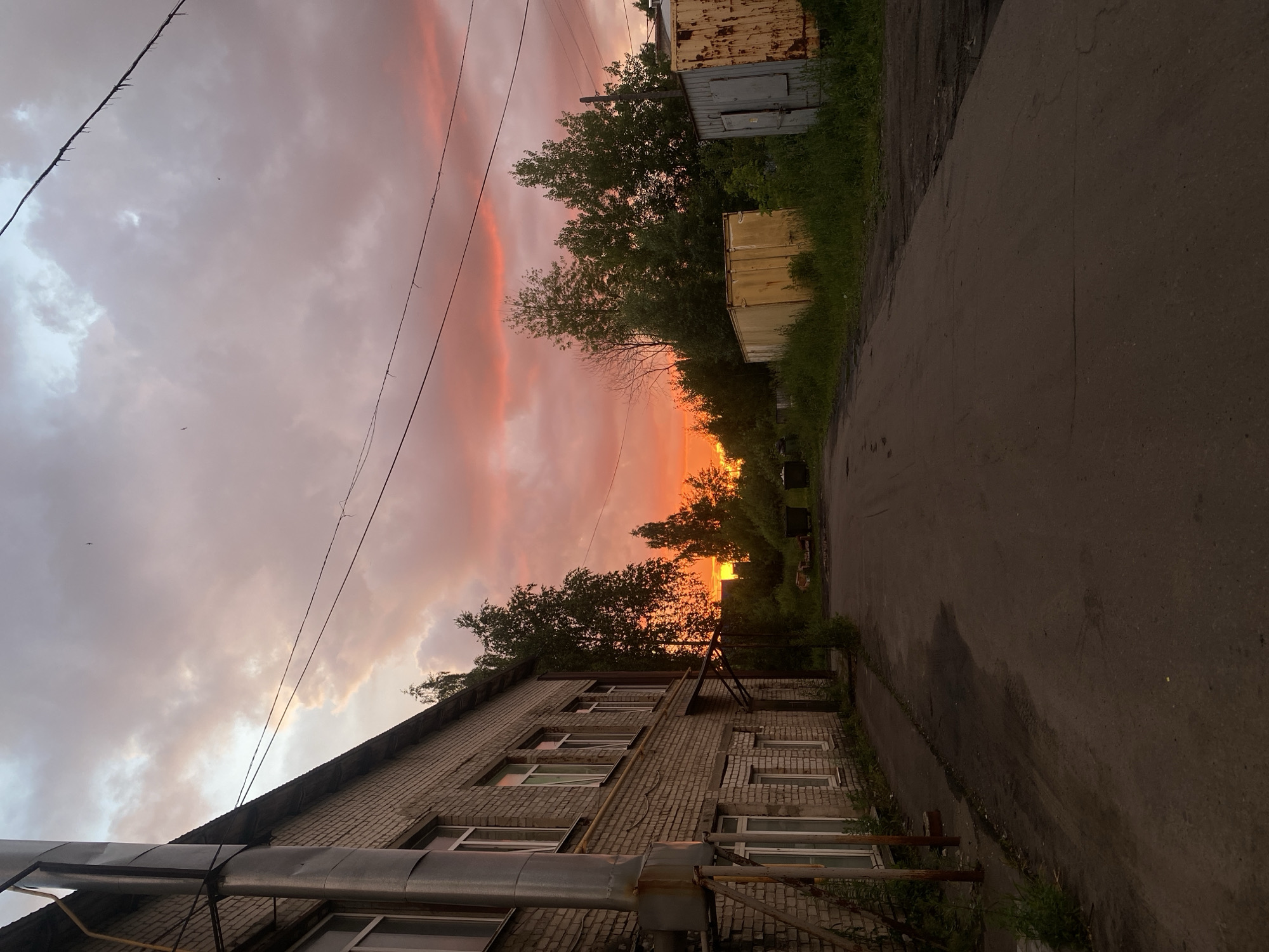 The image shows a street scene at sunset in Dyakovo, Russia. On the left is a brick building with several windows and a metal pipe running vertically along its exterior. The street itself is paved and appears wet in places. To the right of the street, there is overgrown grass, several large metal shipping containers, one of which is yellow and rusted, and another is grey with a closed door. Beyond the containers, trees are visible, and the sky is filled with dramatic clouds illuminated by the setting sun, casting a pink and orange glow. The overall impression is one of an industrial or semi-industrial area during twilight. There are no people visible in the image.