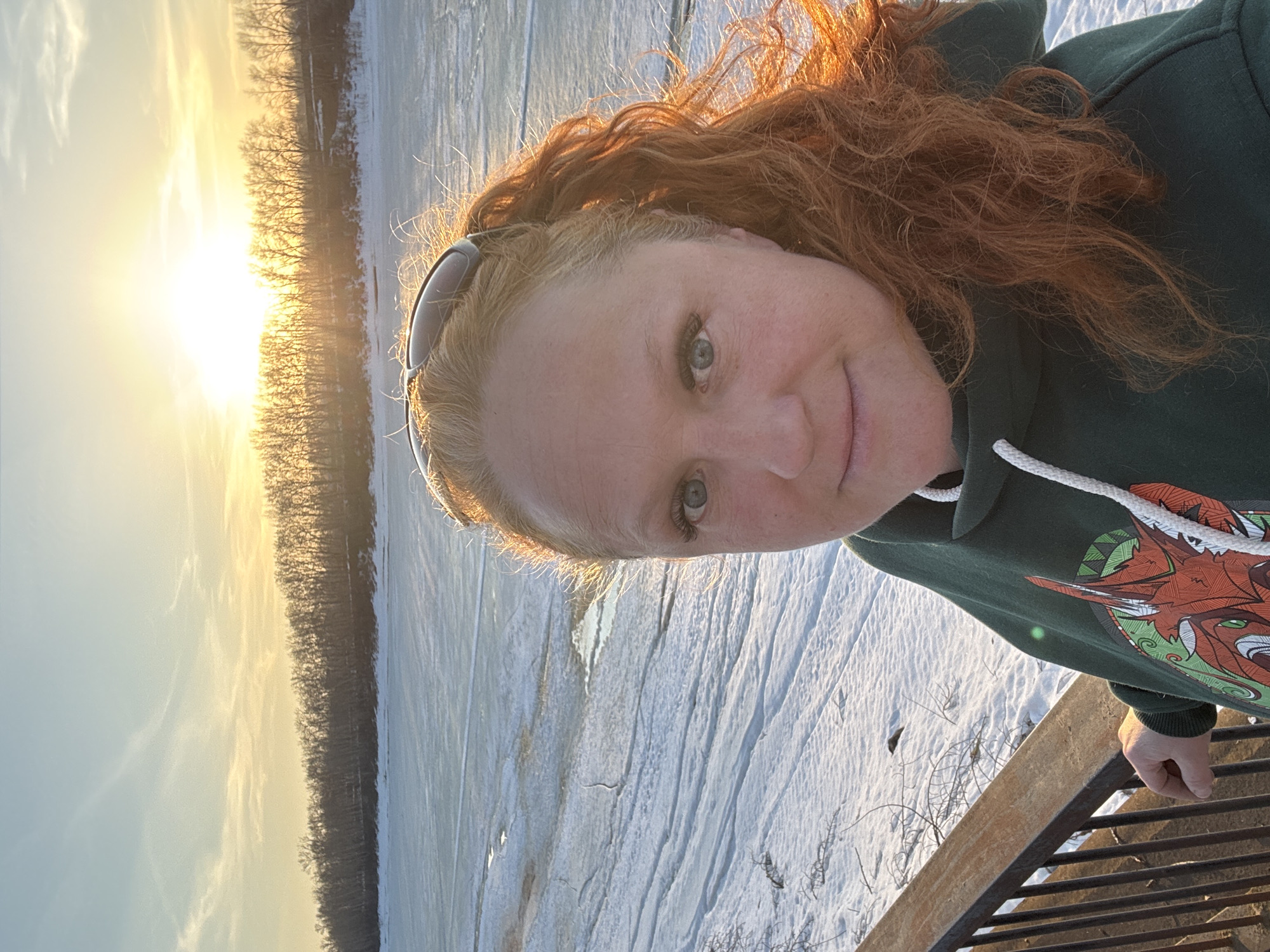 A photograph shows a woman in the foreground and a winter scene in the background, likely taken in the city of Chaikovsky, Russia. The woman has fair skin, blue eyes, and reddish-brown hair, styled up with sunglasses atop her head. She wears a dark green hooded sweatshirt with a patterned graphic, and she appears to be smiling slightly.
The background shows a snowy landscape with bare trees. The sky is bright with the setting sun, casting a golden glow over the scene. A frozen body of water or snow-covered field stretches out behind the trees. The photograph seems to be taken from an elevated position, as a metal railing is partially visible in the lower right corner.