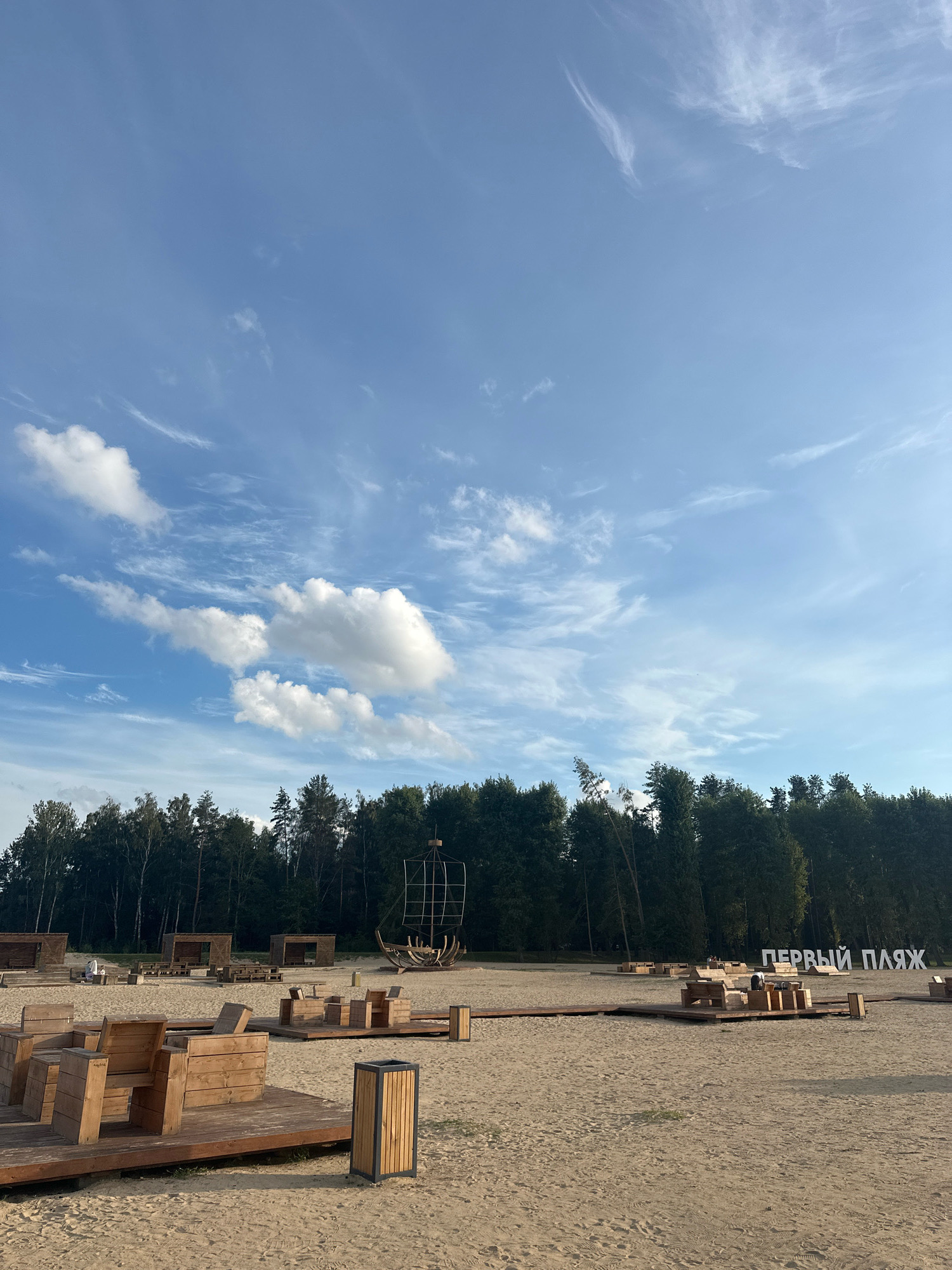 This image depicts a sandy beach area with wooden structures for seating and shade, situated at "Первый пляж" (First Beach). In the background, a line of tall green trees forms a dense forest. To the right, large white letters spell out "ПЕРВЫЙ ПЛЯЖ". A rustic, ship-like wooden structure is visible near the trees. There are several people scattered throughout the beach, some sitting on the wooden furniture, and others walking or standing. The sky above is a clear blue with scattered white clouds. The overall scene suggests a public recreational area designed for relaxation and leisure by a wooded landscape.
