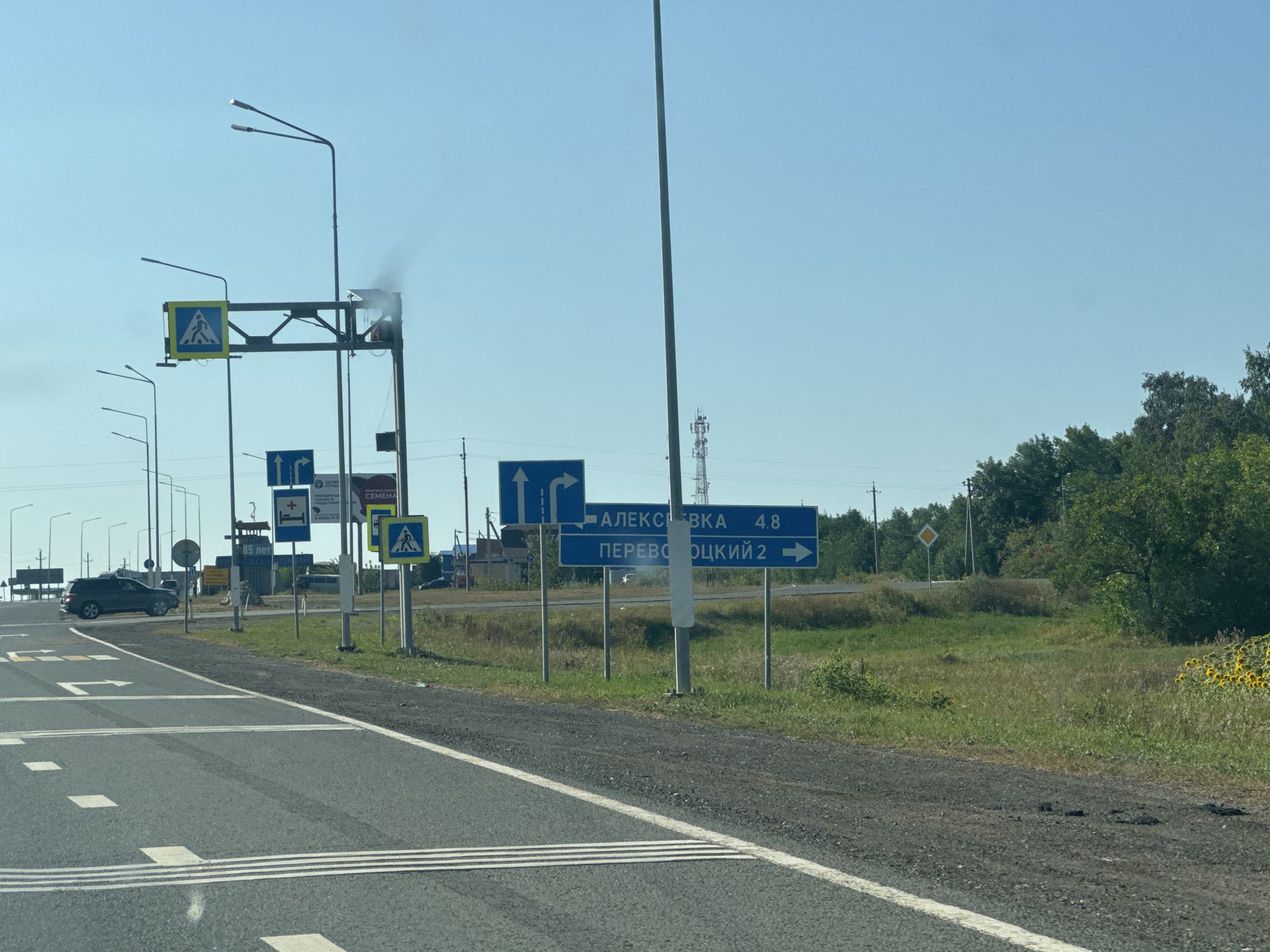 The image captures a roadside scene near Perevolotsky, Russia. In the foreground, the asphalt road features white lane markings and textured tactile paving for visually impaired pedestrians. To the left, a dark-colored SUV is parked on the shoulder. Further down the road, multiple traffic lights and poles are visible. A prominent blue road sign indicates distances: "АЛЕКСЕВКА 4.8" (Alexeevka 4.8) and "ПЕРЕВОЛОЦКИЙ 2" (Perevolotsky 2) with arrows pointing to the right. Another blue sign depicts a right turn. Overhead, a pedestrian crossing sign with a yellow border is mounted on a gantry. In the background, a cell tower stands tall, and lush green trees line the horizon under a clear blue sky. Some sunflowers are visible on the right side of the road. A small amount of smoke or steam is emanating from a structure on the gantry.