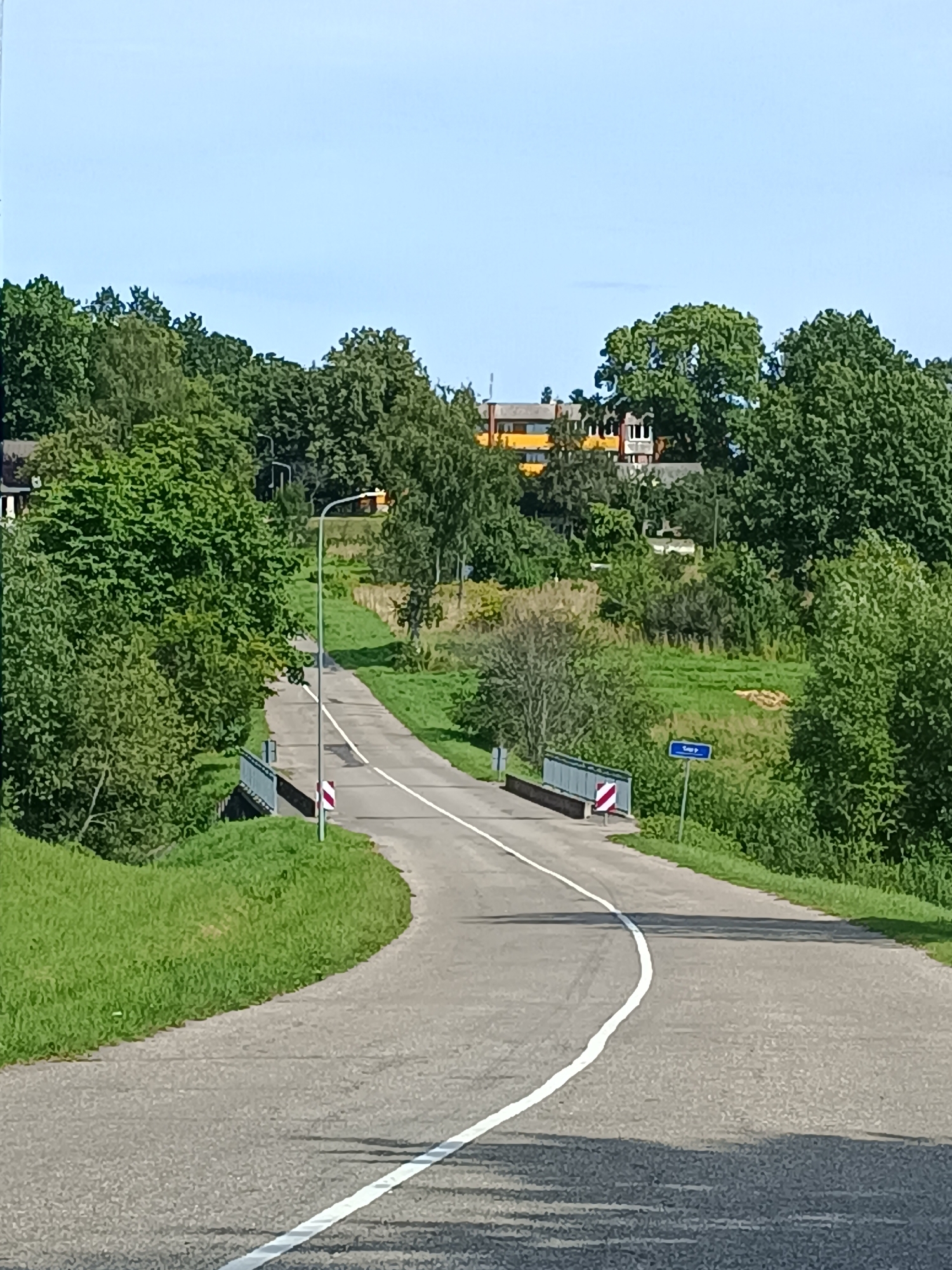 The image depicts a rural road in Aizpute, Latvia, on a clear day under a bright blue sky. The road curves to the right, bordered by lush green grass and dense trees. A small bridge crosses a ditch or small waterway on the left side of the road, with a metal railing and a red and white striped marker. Another red and white striped marker is visible further down the road on the right. A blue road sign with white text, possibly indicating a place name, is situated to the right of the road near the bridge. In the background, several buildings are visible amongst the trees, including what appears to be an orange-colored building. The overall scene is peaceful and serene, characteristic of the Latvian countryside.