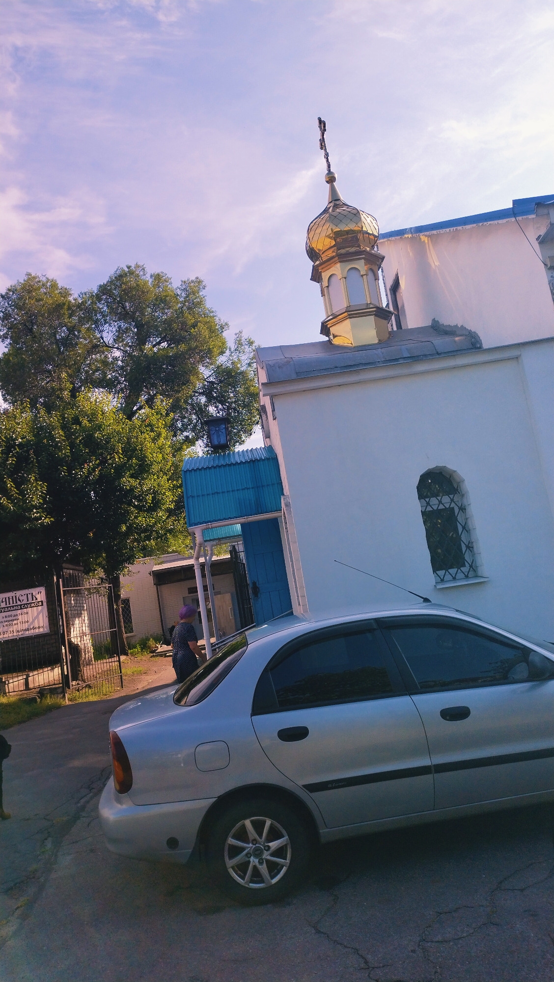 A silver sedan is parked on a cracked asphalt surface in front of a white church with a golden dome and a blue roof. A woman in a polka-dot dress and a purple headscarf stands near a blue entrance to the church. A large green tree is visible to the left of the church, and a fence with a sign is in the background. The sky is partly cloudy with a pink and blue hue. The scene appears to be in Horishni Plavni, Ukraine.