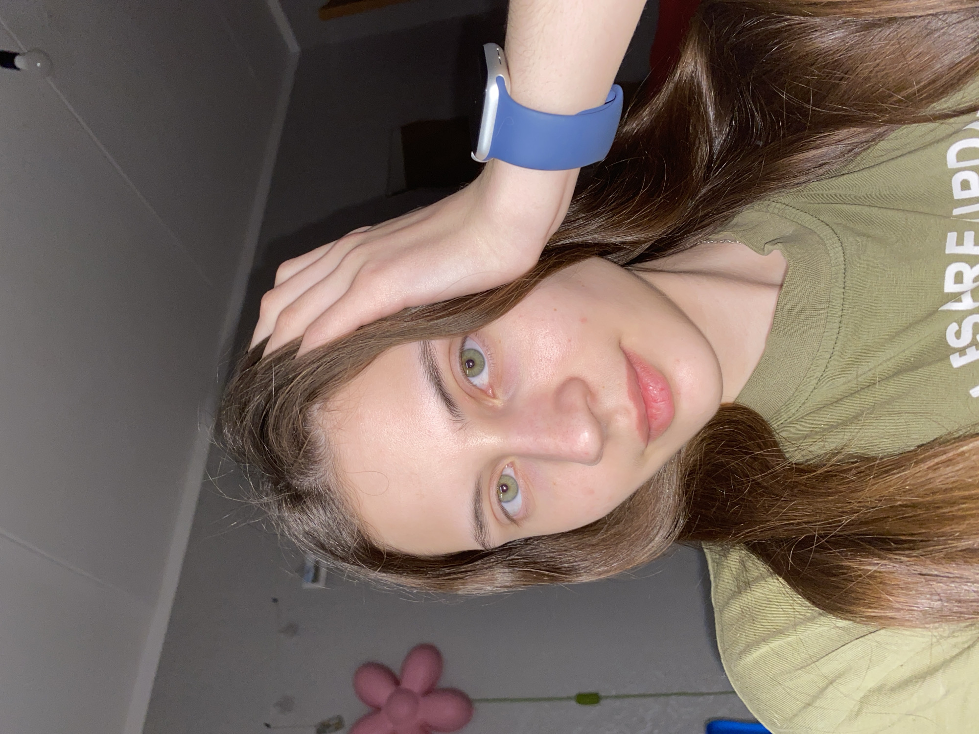 A selfie captures a young person, likely female, indoors with dim lighting. She has long, flowing dark brown hair, fair skin, and prominent green eyes, looking directly at the camera with a neutral to slightly smiling expression. She is wearing an olive-green short-sleeved t-shirt, with white text partially visible on the right side, appearing to read "ESRPEARD" or similar. Her left hand is raised, resting on her head, and she wears a smartwatch with a silver casing and a distinctive blue band on her wrist.
The background is simple and somewhat dark, featuring a light-colored ceiling with a single exposed light bulb fixture visible in the upper left corner. A dark wall or surface is behind her, and in the lower left, a pink, flower-shaped soft object or toy rests on a grey floor. The artificial lighting suggests the photograph was taken in the evening or at night. The setting appears to be a casual indoor space, possibly a home or bedroom, consistent with a personal photograph taken in Votkinsk, Russia, as per the provided context.