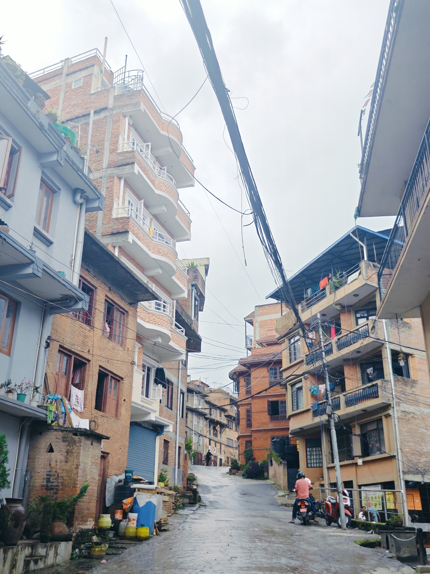 A narrow, wet, paved street winds uphill through a dense urban area in Kirtipur, Nepal, lined with multi-story brick and concrete buildings. The buildings, varying in height and architectural style, feature balconies, numerous windows, and external electrical wiring. Some buildings have laundry hanging out to dry, and potted plants are visible on ledges and in front of some structures. In the foreground, on the left side of the street, are several large ceramic pots, plants, and containers such as buckets and canisters. A motorcycle with a rider wearing an orange shirt is moving uphill on the right side of the street, with another scooter parked behind it. Further up the street, a person is visible on a bicycle. The sky is overcast.