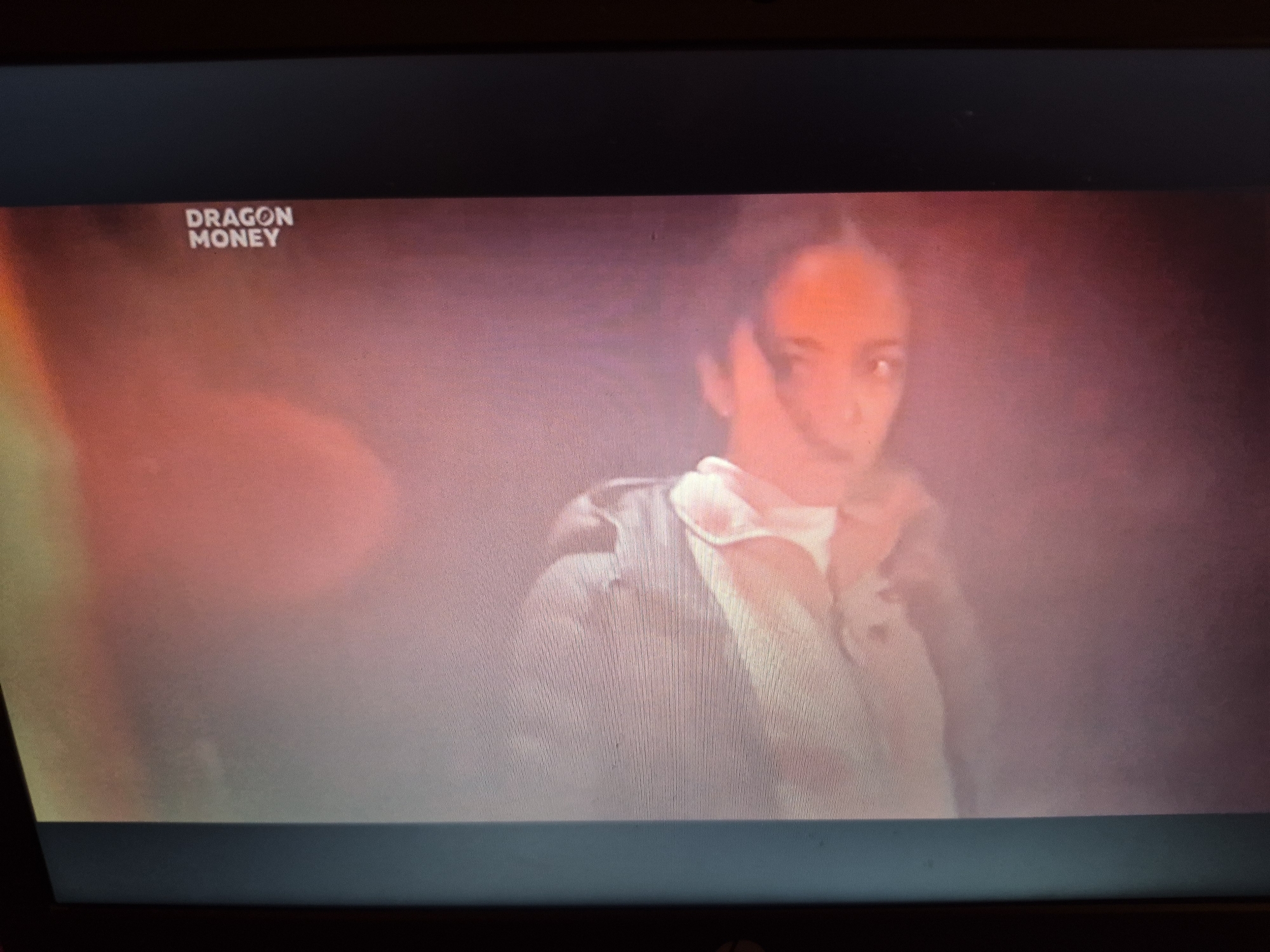 The image shows a television screen displaying a scene with the text "DRAGON MONEY" in the upper left corner. A person, likely a woman, is visible in the center right of the screen, appearing to be wearing a light-colored jacket or uniform. The background is dark and hazy with reddish and pinkish tones, suggesting a dramatic or atmospheric setting. The image is a photograph of a screen, with some glare and digital artifacts visible. There is no indication of the location being Sandwell, United Kingdom.