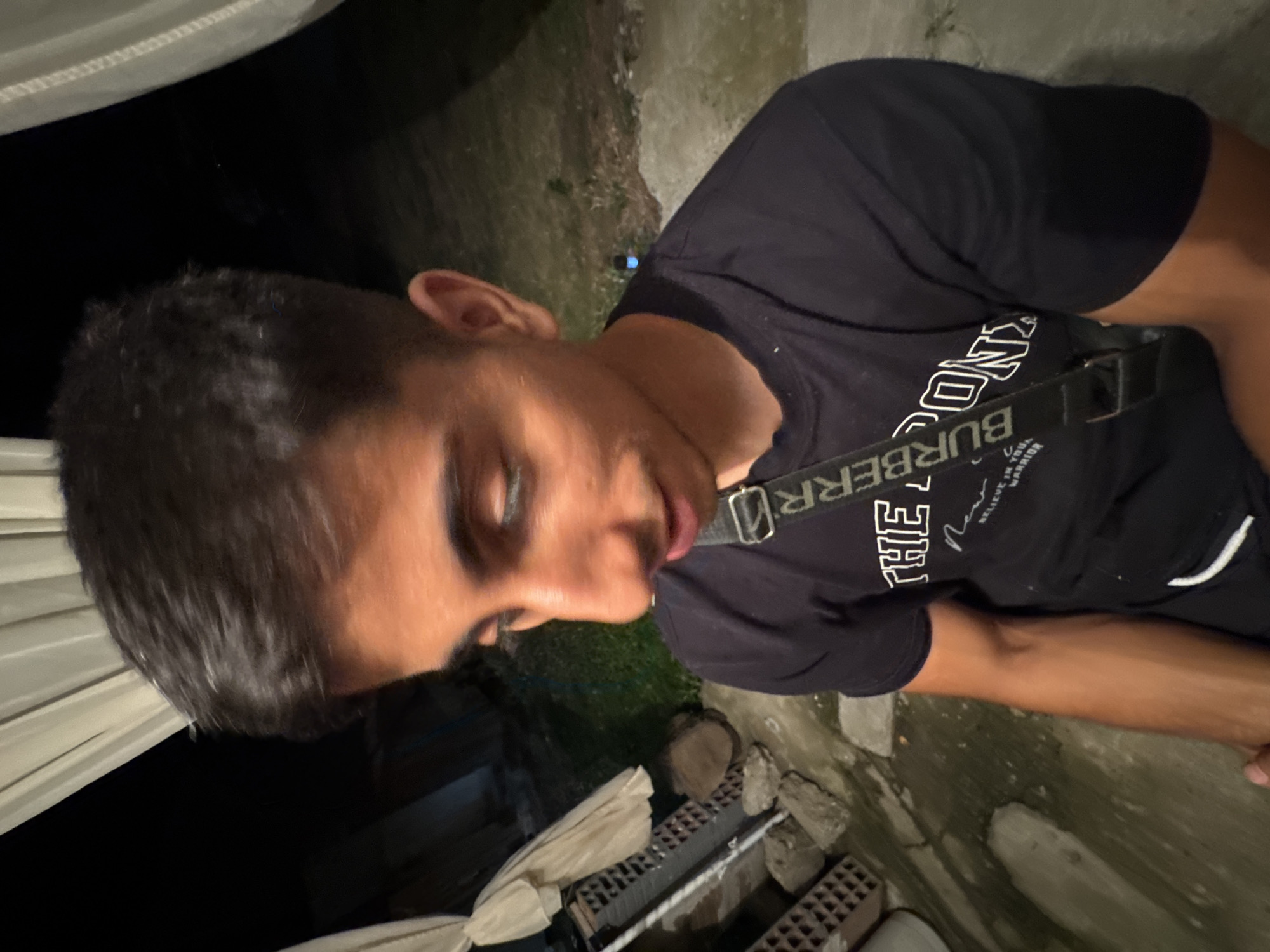 The image shows a young man with dark hair and brown skin, wearing a black t-shirt with a printed design and a black strap across his chest. He is looking down and slightly to the side, with his mouth open. The background is dark and appears to be an outdoor setting with some draped fabric in the upper left corner and what looks like stone or concrete ground. There are also some piles of bricks and rocks visible. The overall lighting suggests it is nighttime. The scene is not specifically identifiable as being in the city of Sredets, Bulgaria.