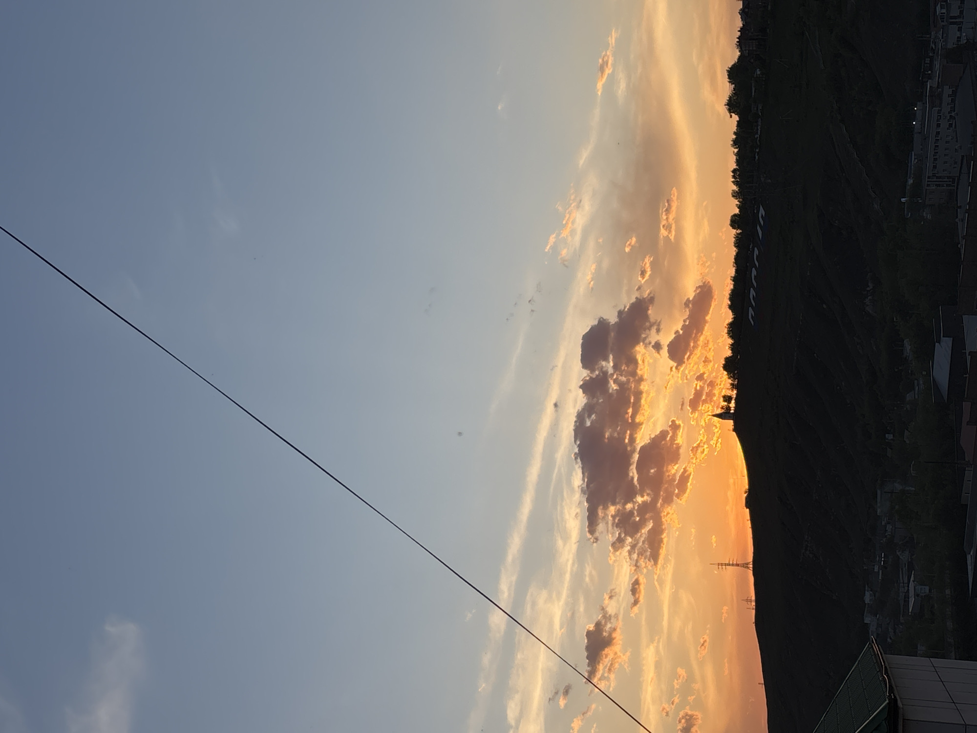 The image shows a landscape with a dramatic sunset sky over Krasnoyarsk, Russia. The sky transitions from a light blue on the left to vibrant oranges and yellows on the right where the sun is setting behind a large, dark hill or mountain. Clouds are scattered throughout the sky, reflecting the warm colors of the sunset. The silhouette of the hill is punctuated by trees and structures, including what appear to be power lines. On the hill, a large word, likely the city's name, is placed on the mountain side. On the bottom right, there's a view of buildings and structures, indicating an urban area. A power line cuts across the image from the top left corner towards the bottom right.