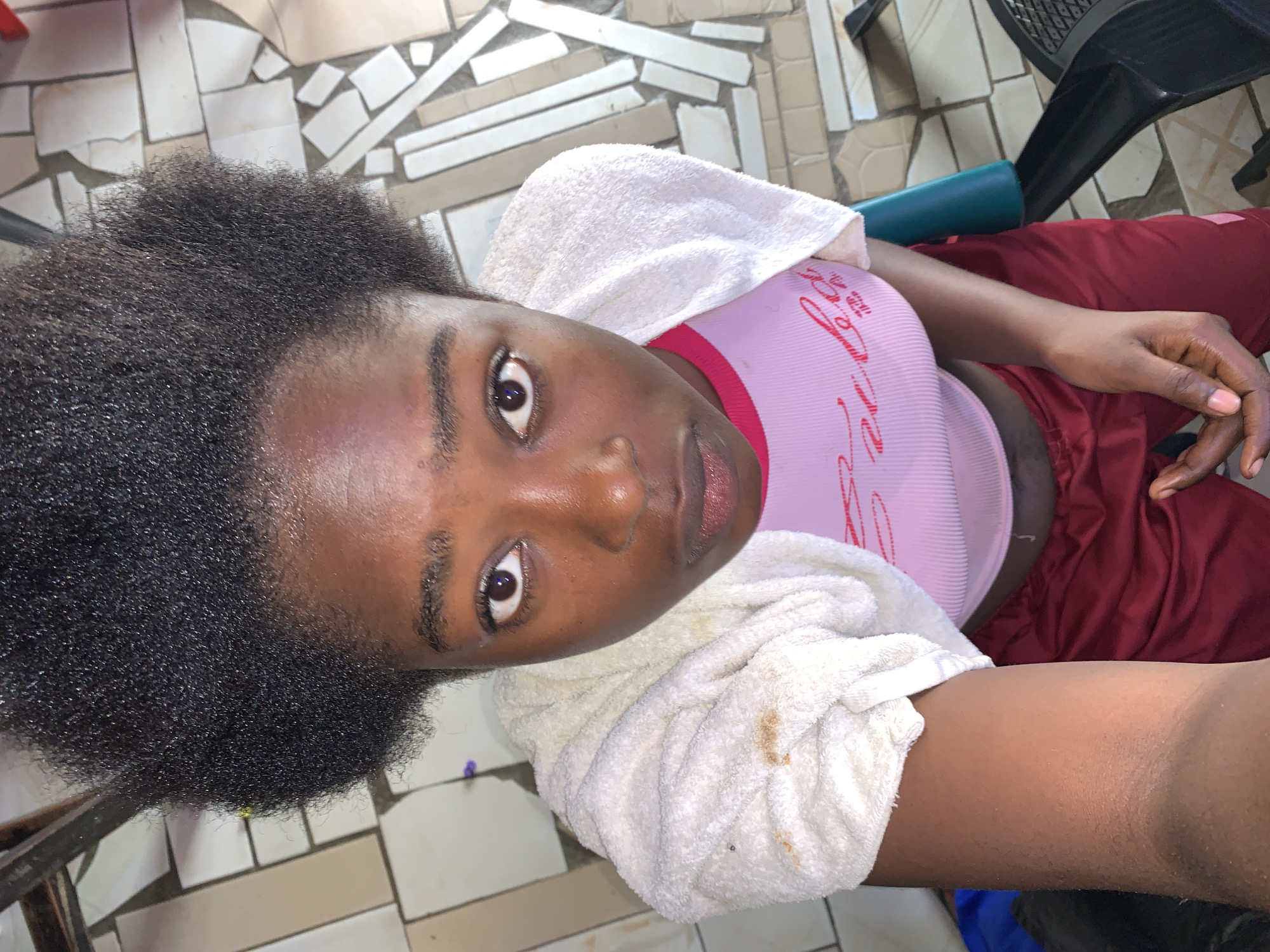 A young woman with dark, natural hair and medium-brown skin lies on a tiled floor. She wears a pink top and maroon sweatpants. A white towel is draped around her shoulders. The background shows a tiled floor in an indoor space. Based on the prompt, the location is Rafin Par, Nigeria.