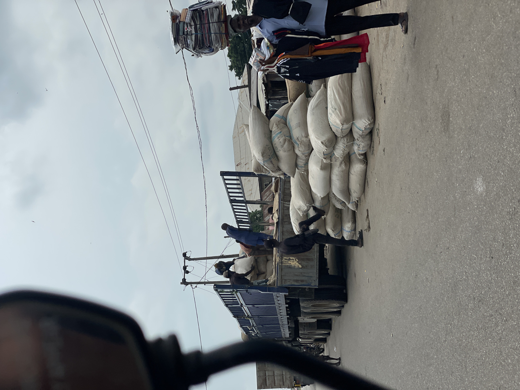 In Lokoja, Nigeria, a street scene unfolds with a large truck filled with sacks, likely agricultural produce, being unloaded. Several individuals are engaged in the laborious task of moving these sacks. One person is in the truck bed, actively involved in the loading or unloading process. Another individual, dressed in a blue outfit, is standing on the ground beside the truck, appearing to assist. A third person, in black attire, is poised near a stack of sacks, one leg raised as if about to step or having just stepped. Further down the street, a vendor carries a substantial load of folded clothing or fabric items stacked high on their head, balanced with remarkable skill. This vendor is also holding an assortment of garments on a hanger, presumably for sale. The environment suggests a bustling market or commercial area, with the sky overcast and overhead electrical wires present. The overall impression is one of a busy, working day in a Nigerian city.