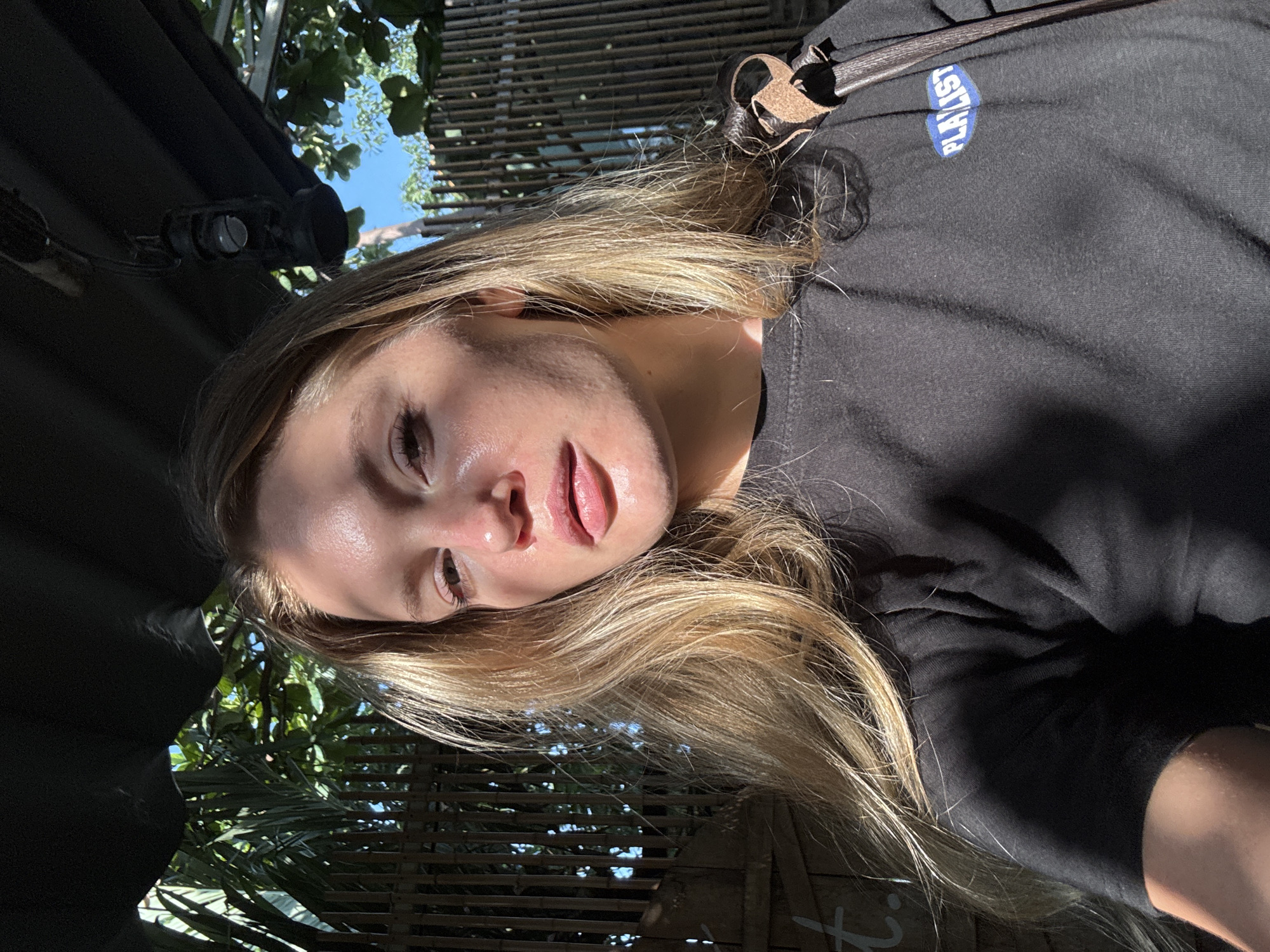 In Nha Trang Ward, Vietnam, a woman with blonde hair, wearing a black t-shirt with a blue and white oval logo, is captured in a close-up, selfie-style portrait. Sunlight illuminates her face, highlighting her features and casting shadows across her skin. She is outdoors, with a backdrop of lush green foliage and a bamboo fence. A dark object, possibly a lantern, is suspended from above. A brown strap, part of a bag, is visible over her shoulder. The overall scene suggests a casual, outdoor setting, possibly a cafe or a resort area, given the tropical vegetation and decorative elements.