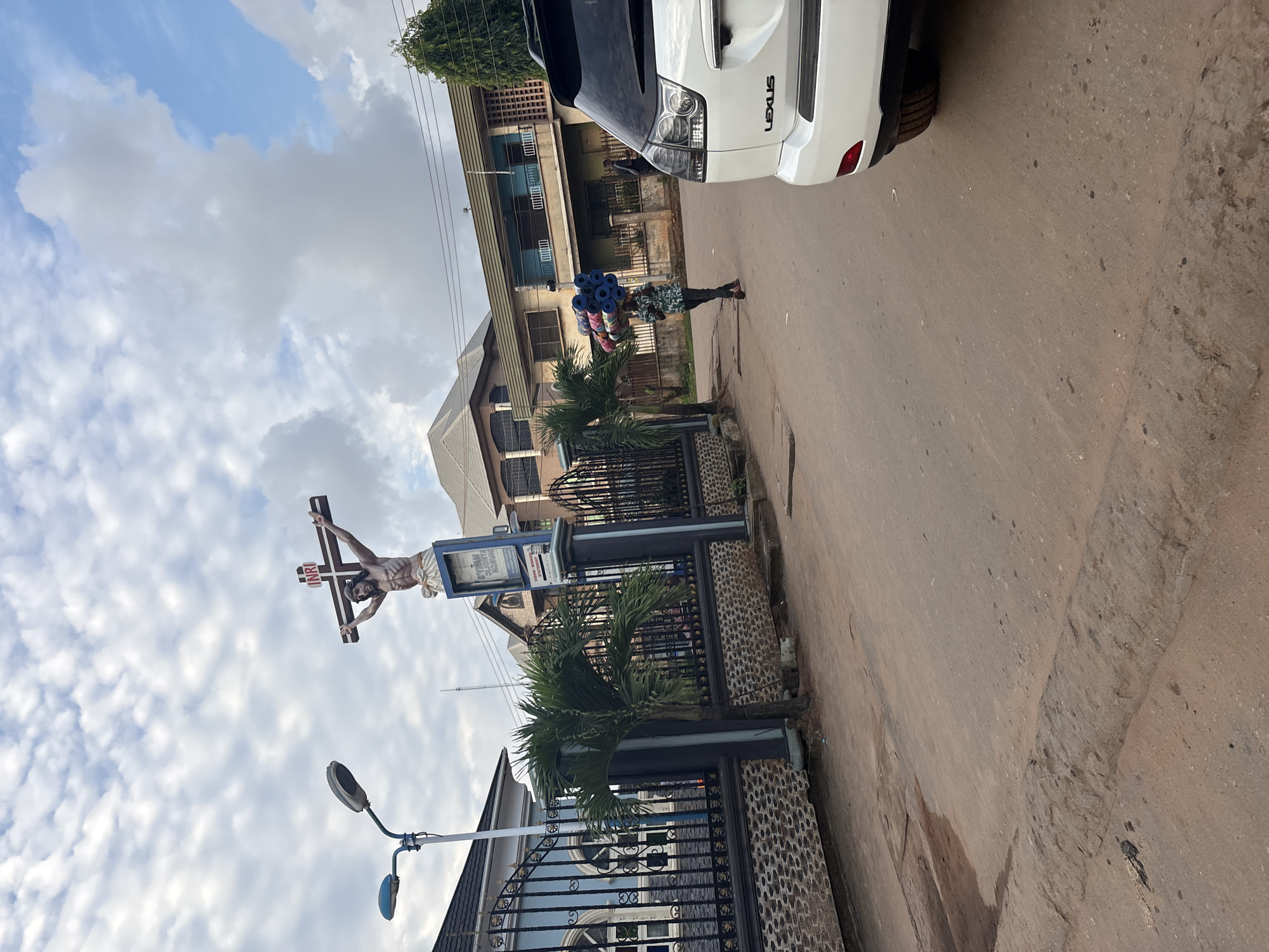 The image depicts a street scene in Onitsha, Nigeria. In the foreground, a white Lexus SUV is parked, partially obscuring a red-brown asphalt road. To the left, a black metal gate with intricate designs and a stone-patterned base leads to a blue building. A lamppost with two lights stands beside the gate. Further back, a prominent statue of Jesus Christ on the cross is visible, with the inscription "INRI" above his head. The statue is placed on a pedestal that includes a blue sign with white text. A palm tree stands in front of the gate and fence. In the middle ground, a person is walking away from the viewer, carrying a stack of rolled-up blue mats on their head. They are wearing a patterned shirt and dark pants. Behind this individual, several multi-story buildings with balconies and tiled roofs line the street. The sky is partly cloudy with patches of blue. The overall impression is of a typical urban environment in Nigeria, with elements of religious iconography and daily life.
