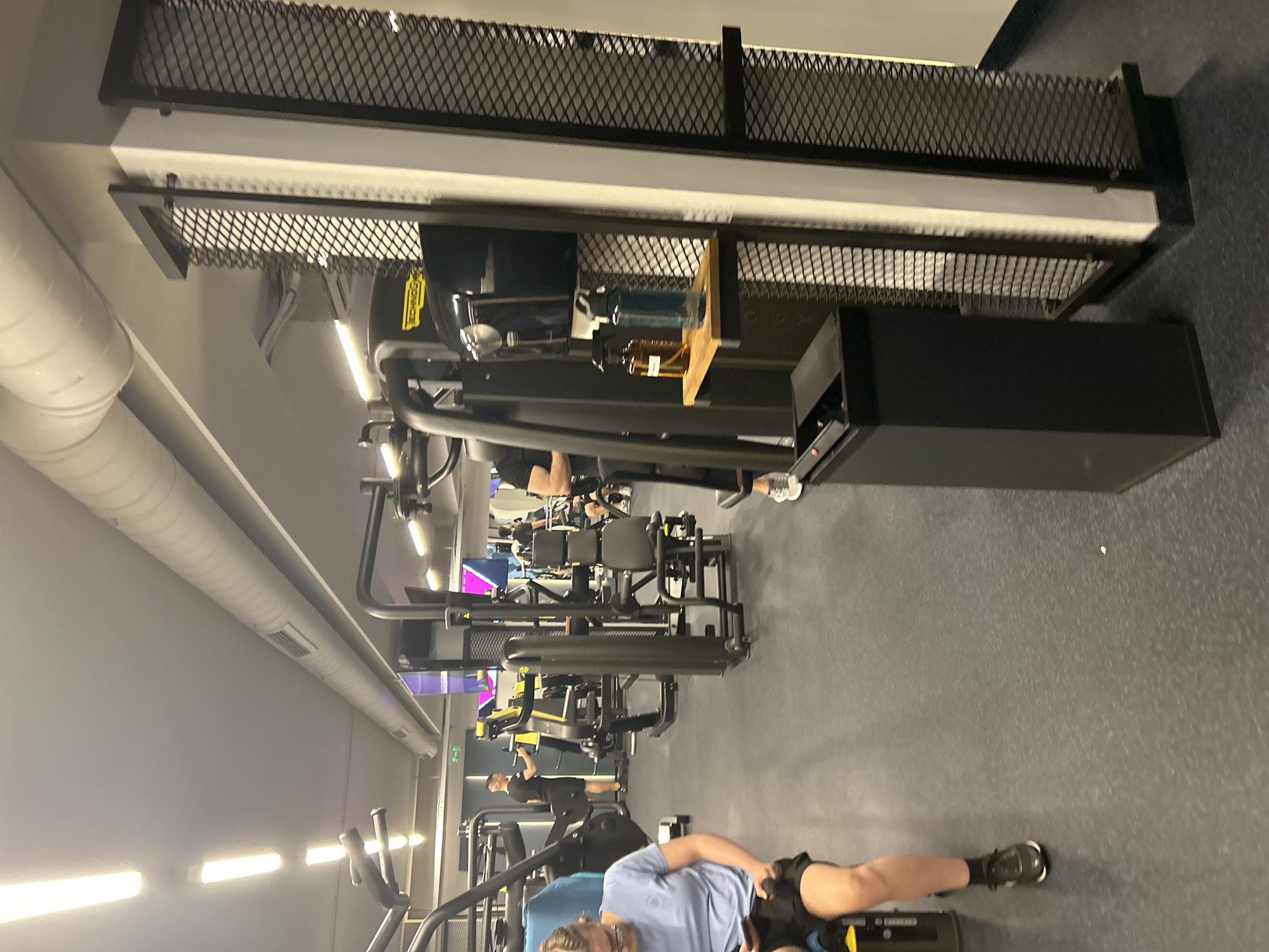 The image shows the interior of a gym, likely in Varna, Bulgaria. Several pieces of exercise equipment are visible, including weight machines and possibly elliptical trainers. People are using some of the equipment. The gym has a grey floor. Lighting appears to be a mix of overhead fluorescent lights and natural light. There's some metal mesh construction overhead.
