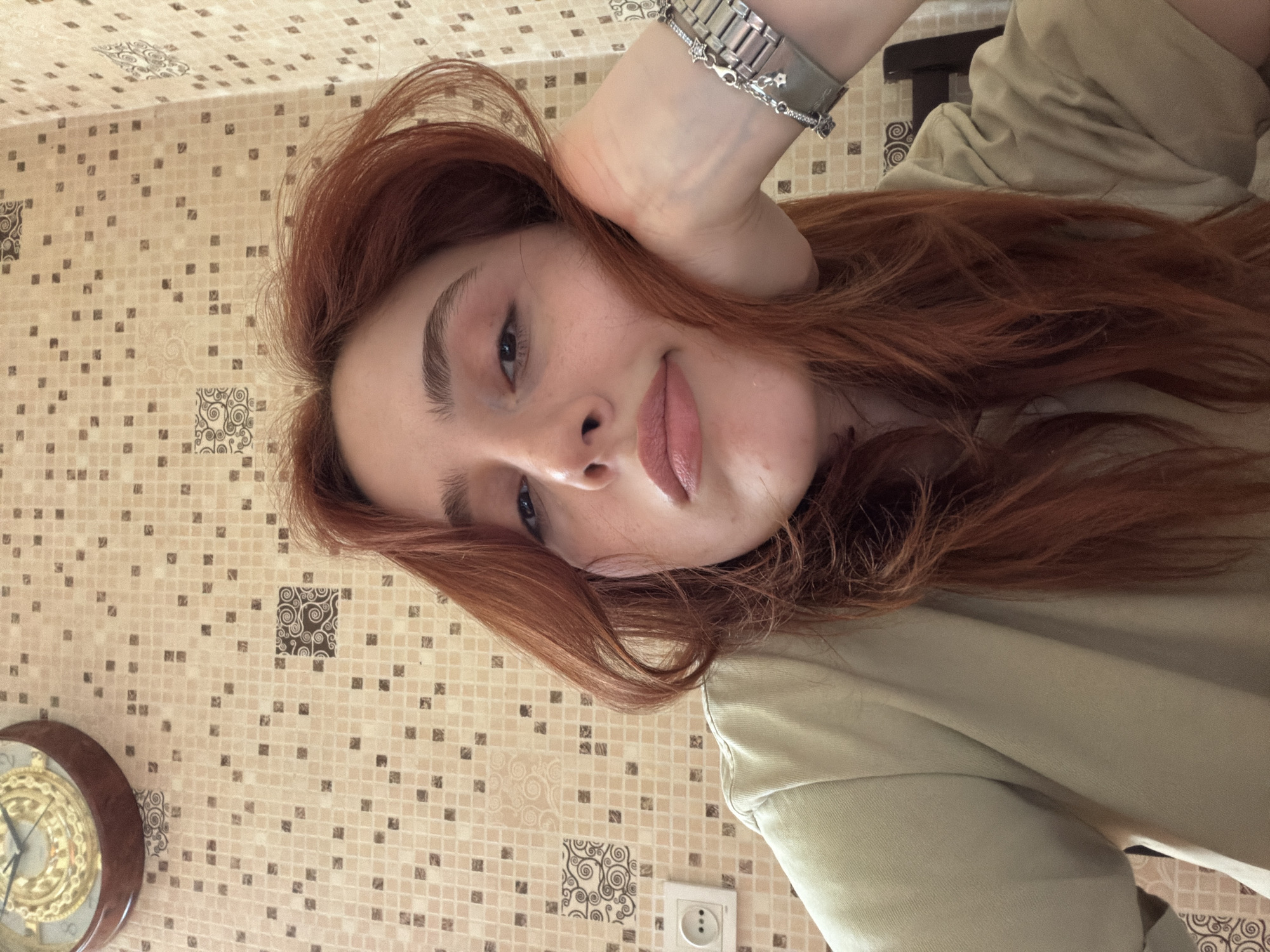 A young woman with long, reddish-brown hair is posing for a photo. Her head is tilted slightly, and she is looking directly at the camera with a small, subtle smile. She is wearing a light green or beige long-sleeved shirt. On her left wrist, she has a silver-colored watch with decorative elements on the bracelet. Her hand is resting on her head, with the bracelet prominently displayed.
The background consists of a tiled wall with a mosaic pattern of small squares in beige and brown tones. There are decorative tiles with swirling patterns scattered throughout the wall. In the lower left corner of the image, there is a partially visible ornate wall clock with a dark brown frame and golden details. In the lower right corner is an electrical outlet.
Without additional information, I cannot determine the specific location within the city of Tbilisi, Georgia.