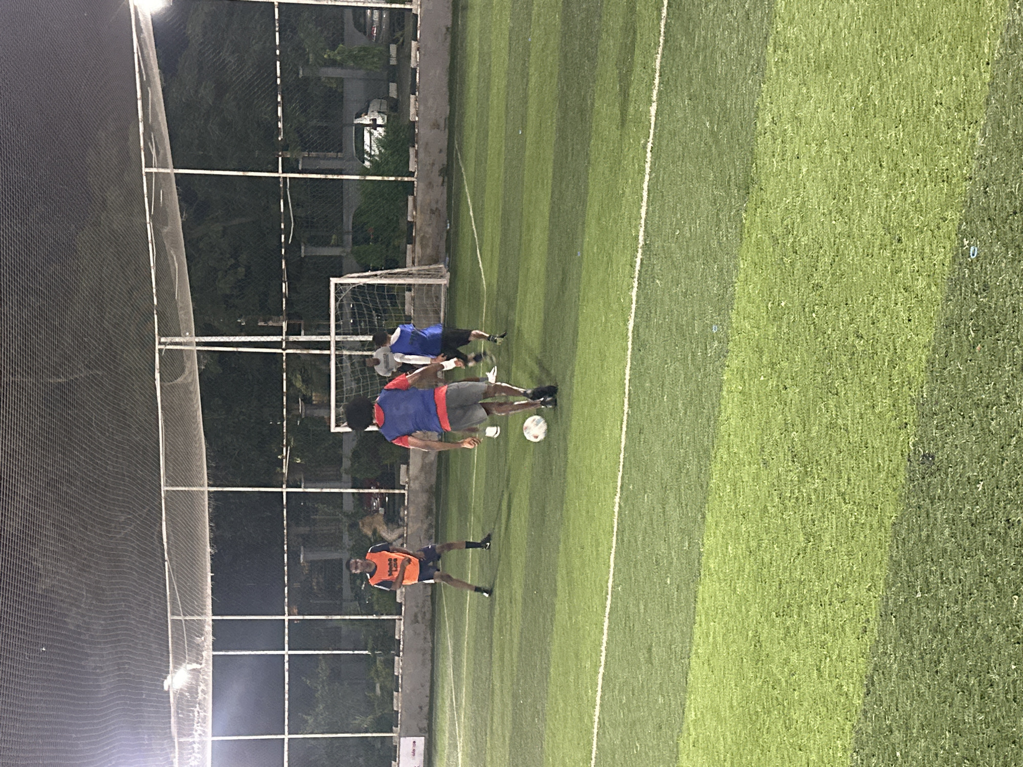 In the city of Abuja, Nigeria, on an outdoor, artificially turfed football pitch, three individuals are engaged in a football match. The pitch is marked with white lines. A net enclosure surrounds the playing area, and lighting fixtures illuminate the field from above. In the background, trees and parked vehicles are visible, suggesting an urban setting. Two players are wearing vests over their shirts, one orange and one blue, and the third player is in a blue vest and dark shorts. The person in the foreground, facing away from the camera, is dribbling a soccer ball. The scene captures a moment of active recreation on a sports field.