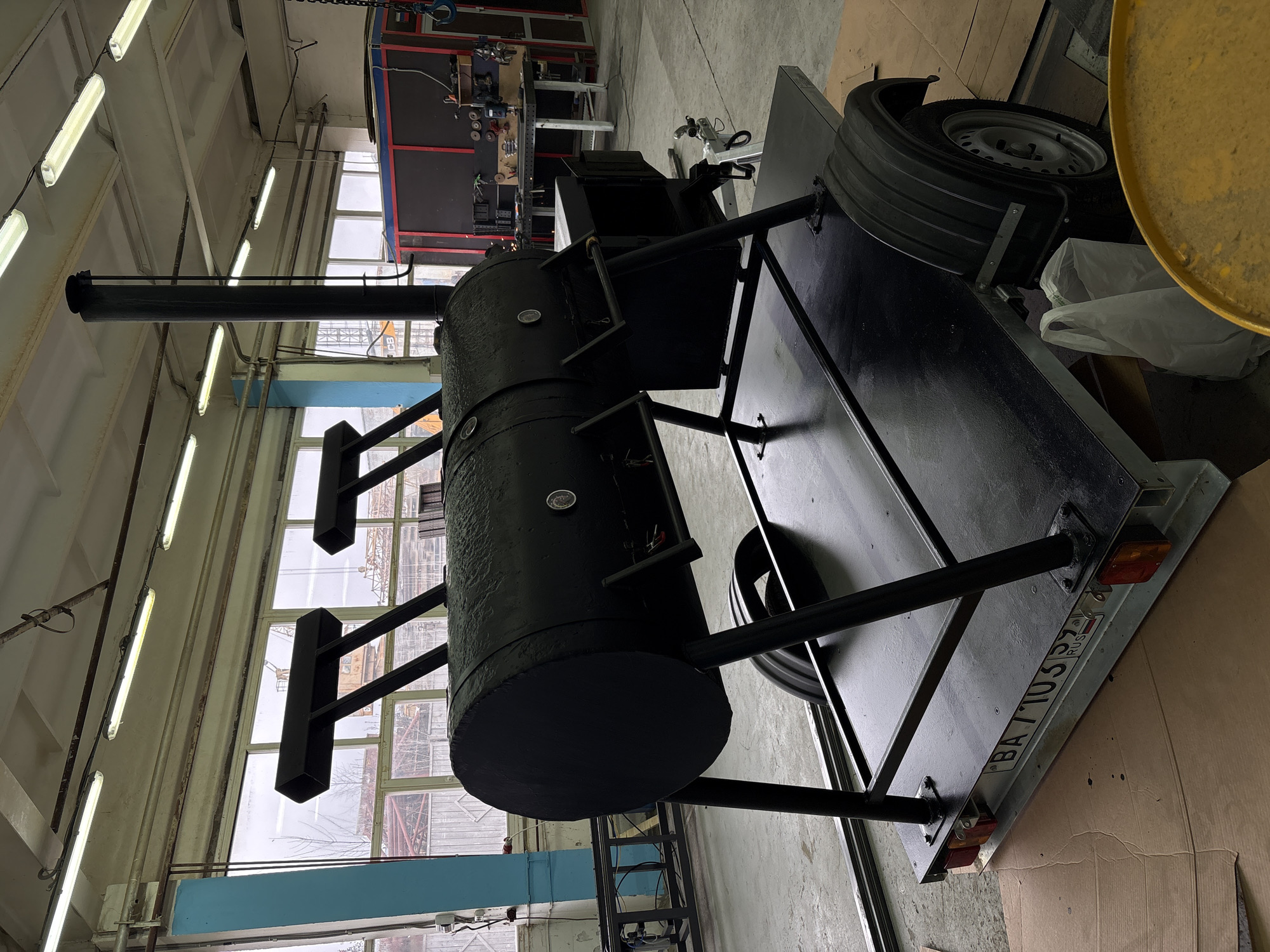 This media file captures a large, black, industrial-style BBQ smoker mounted on a trailer, situated within an indoor workshop or garage. The smoker is a prominent cylindrical unit with multiple hatches, handles, and embedded thermometers, featuring a tall, dark smokestack extending upwards. It is securely affixed to a black flatbed trailer, which also holds a spare tire on its side, partially obscured by a yellow container.
The setting appears to be a manufacturing or repair facility in Chaikovsky, Russia, characterized by high ceilings, a concrete floor, and rows of fluorescent lighting fixtures. Large windows along the upper walls provide natural light and offer a glimpse of an overcast sky and distant industrial structures, possibly cranes. In the background, workbenches and shelves stocked with various tools and equipment are visible, reinforcing the industrial environment.
No individuals are present in the image, and no specific activity is occurring; the smoker and trailer appear to be a completed assembly. The lighting suggests it is daytime. A notable detail is the license plate at the rear of the trailer, which displays the text "BA 7105 | 59 | RUS". The "RUS" clearly indicates Russia, and the "59" is the regional code for Perm Krai, confirming the provided location context of Chaikovsky.