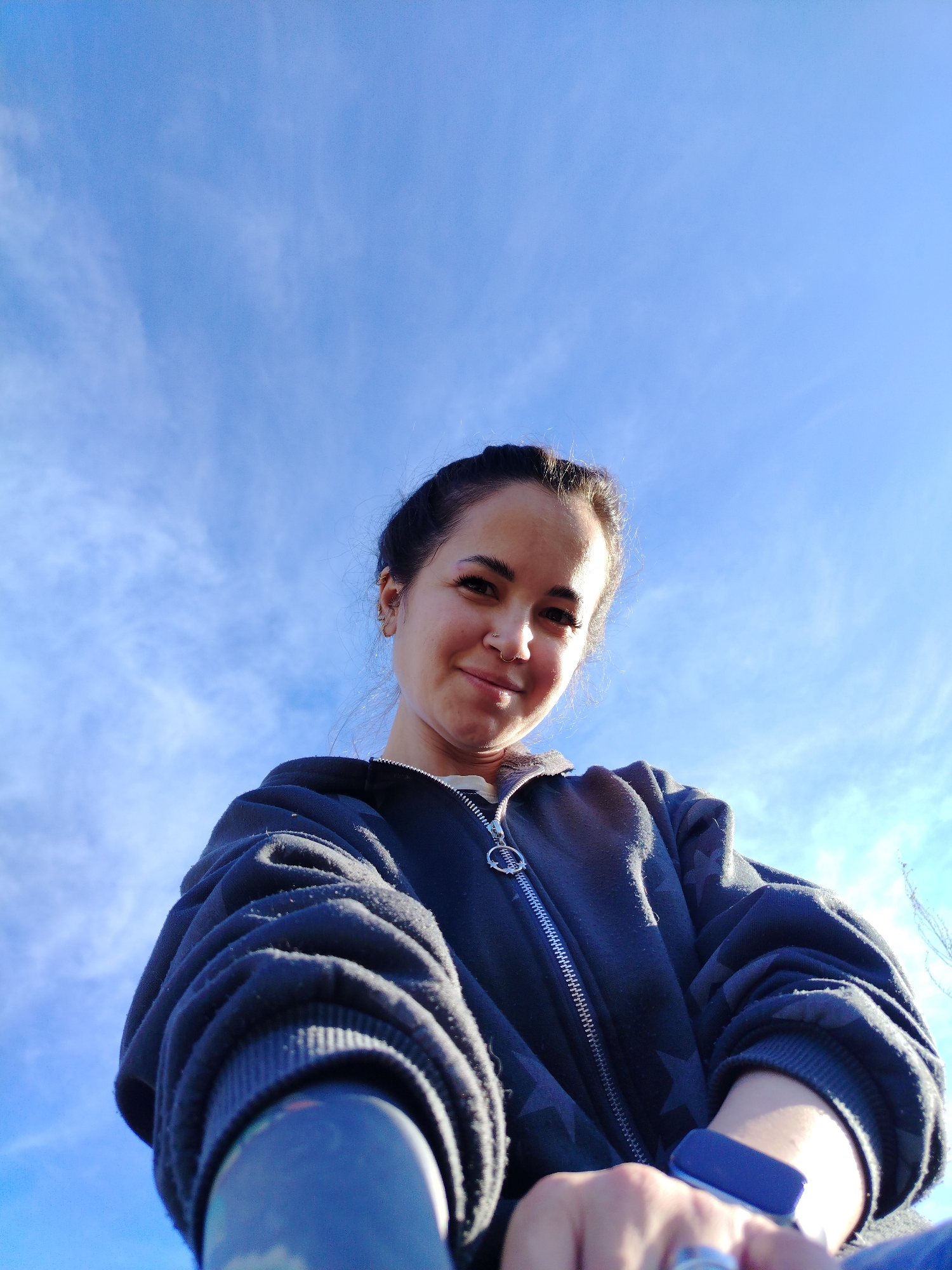 A young woman poses for a low-angle selfie against a bright blue sky with wispy clouds. She is wearing a dark zip-up hoodie with star patterns. A blue fitness tracker is visible on her wrist. She has her hair pulled back and is wearing a nose ring and multiple earrings. The photograph appears to have been taken outdoors. There is no verifiable information to suggest that the picture was taken in the city of Гожан, Russia.