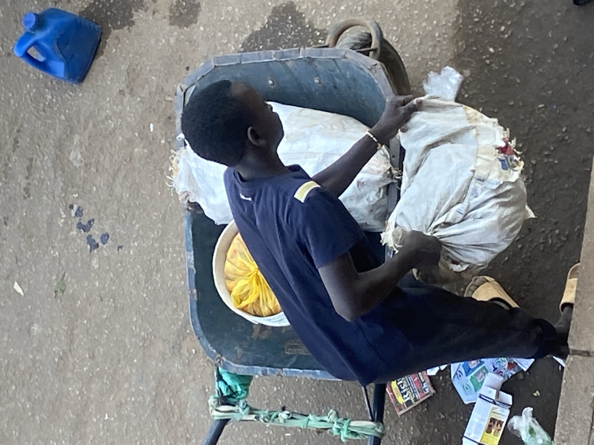 The image shows a young person, likely male, wearing a dark blue t-shirt and dark pants, standing next to a wheelbarrow filled with various items. The wheelbarrow contains at least two large white sacks and a yellow bag. The person is reaching into the wheelbarrow, appearing to be interacting with one of the white sacks.
The ground around the wheelbarrow is rough and textured, resembling a paved or dirt surface. Scattered on the ground are some discarded items, including what appear to be cardboard boxes, plastic bags, and possibly some packaging materials. To the upper right of the scene, a bright blue plastic container with a handle is visible.
The overall lighting suggests it is daytime, with shadows present on the ground. The perspective is from an elevated viewpoint, looking down onto the scene. The location in Bukuru, Nigeria, is indicated by external information not present in the image itself.