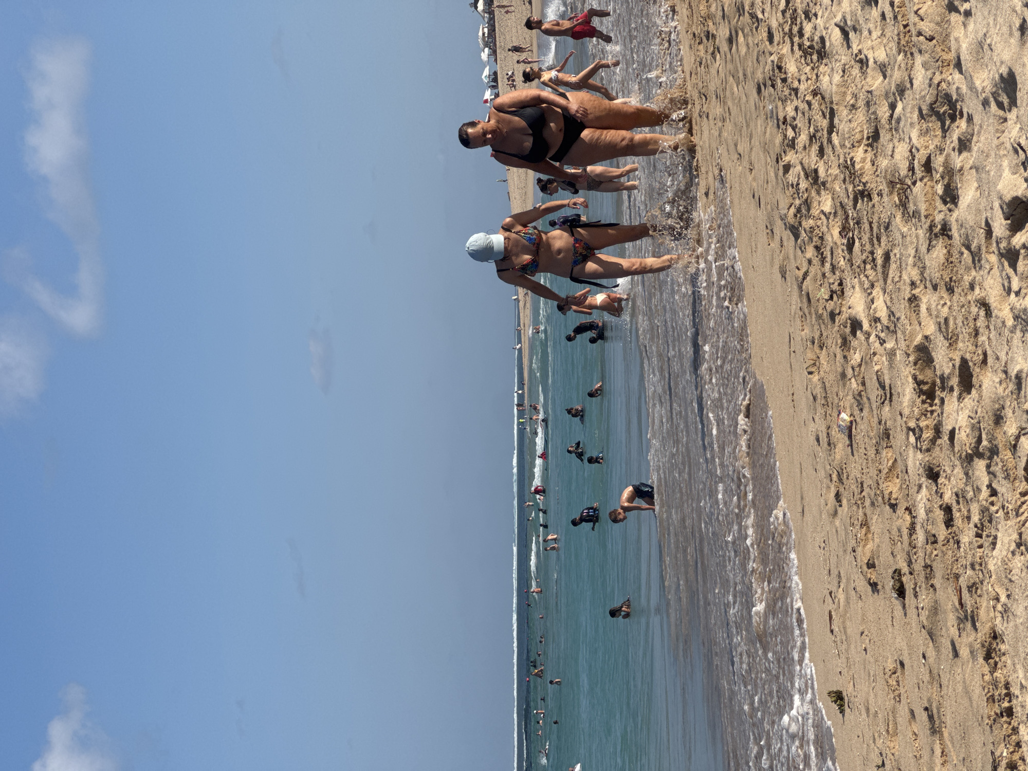 Two women walk through shallow ocean water on a sandy beach, with waves crashing around their ankles. Both are wearing bikinis. The woman on the left wears a colorful patterned bikini and a light blue baseball cap. The woman on the right wears a black bikini. In the background, numerous other people are in the ocean, some wading, some swimming, and some appearing to be children. The ocean is turquoise and calm, with a visible horizon line where it meets a pale blue sky with scattered white clouds. The beach in the foreground is golden sand, with footprints and some debris scattered on its surface. In the far distance, umbrellas and tents suggest a developed resort area. This scene is consistent with a public beach in Kuta Selatan, Indonesia, a popular tourist destination known for its beaches and ocean activities.