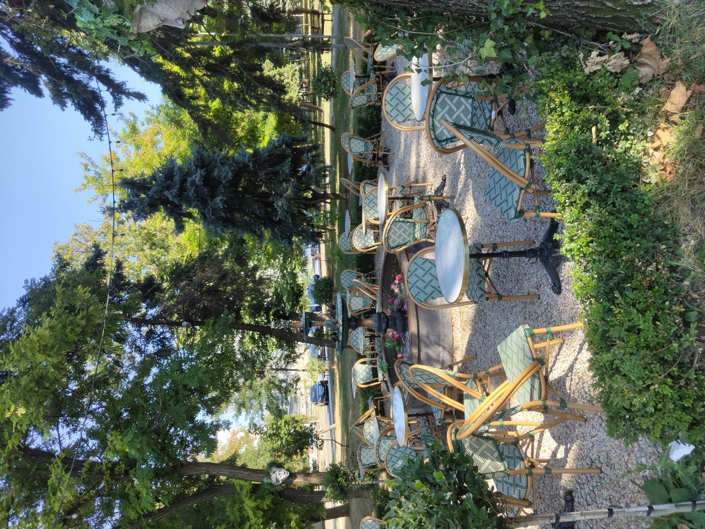 The image depicts an outdoor cafe with multiple tables and chairs arranged on a gravel surface. The cafe is situated in a park-like setting with numerous trees providing shade. In the background, a fountain is visible, and beyond that, cars are parked on a street, suggesting an urban environment. The tables are round and topped with white surfaces, supported by dark metal bases. The chairs have light-colored wooden frames and green patterned upholstery. The overall impression is one of a serene, European-style outdoor seating area. Based on the architectural style of the fountain and the general ambiance, this location is likely a cafe in Chișinău, Moldova. No people are visible in the image.