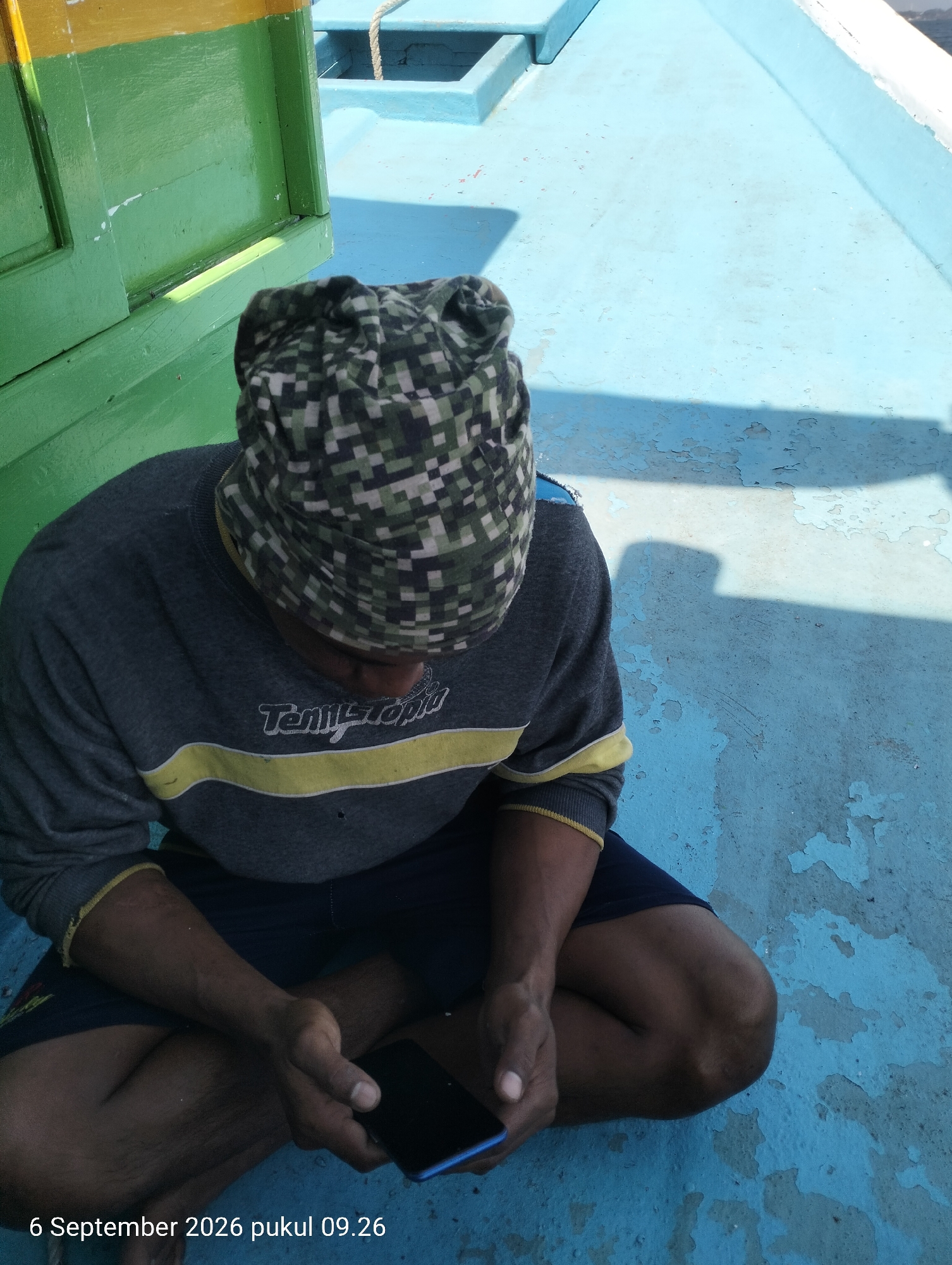 A person wearing a camouflage beanie and a grey long-sleeved shirt with yellow stripes is sitting on a blue surface, possibly a boat deck. The person is holding a dark-colored smartphone in their right hand. The person is looking down at the phone. The background shows parts of a green and yellow structure, a blue water body, and a rope. The scene appears to be outdoors, under sunlight, as indicated by the shadows. The timestamp "6 September 2026 pukul 09.26" suggests the image was taken in Kendari, Indonesia.
