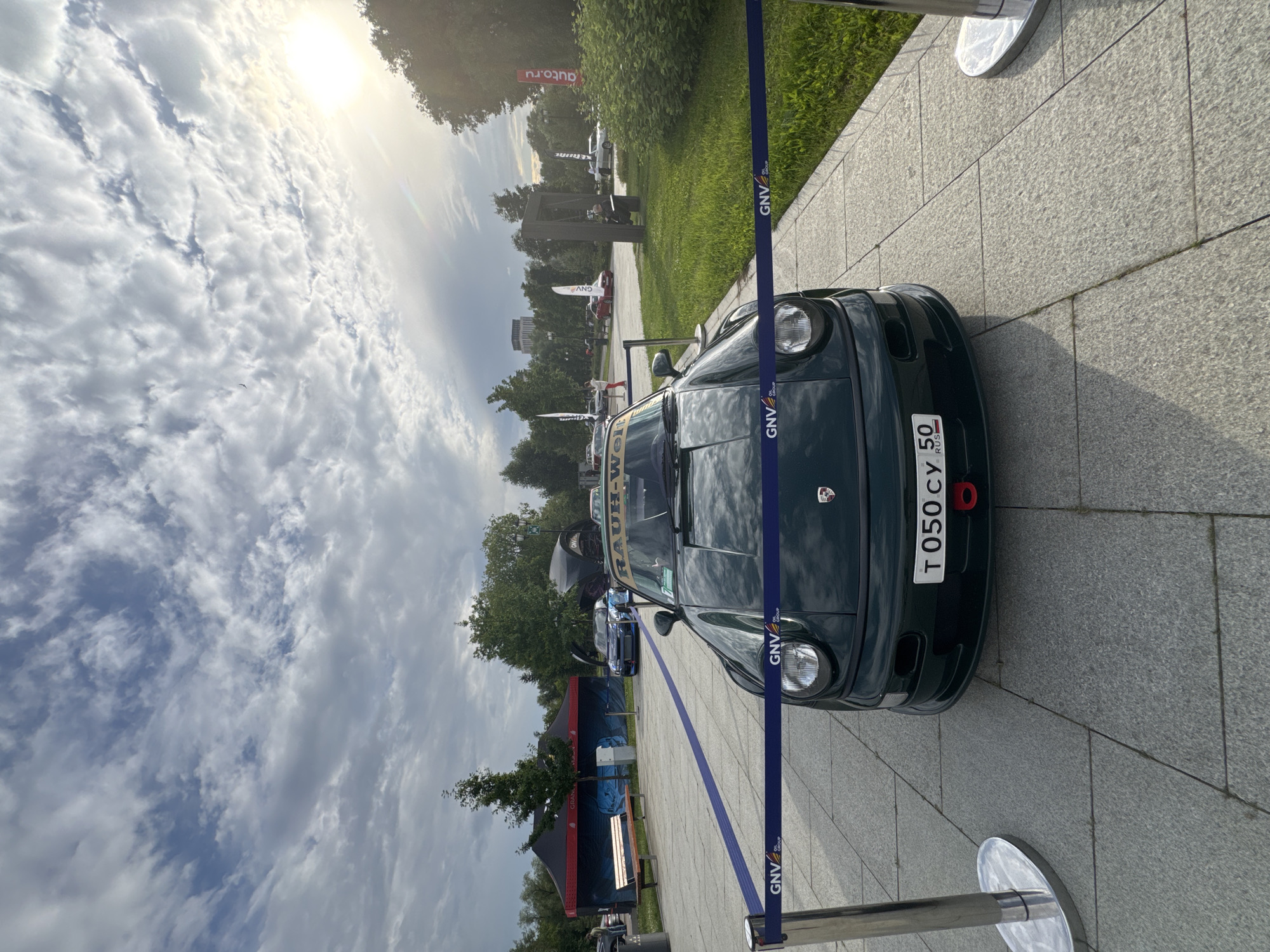 This media file captures an outdoor event, likely a car show or exhibition, set on a paved area with a grassy verge and trees in the background. The sun is bright, casting long shadows, and the sky is filled with dramatic, wispy clouds, suggesting either early morning or late afternoon.
The main subject is a dark green car, possibly a Porsche, with its front clearly visible. It has a license plate that reads "T 050 CY 50" and "RUS". A yellow banner with black text, which appears to be "RAUH-WELT", is affixed to the car. The car is cordoned off by a blue ribbon with "GNV OIL GROUP" printed on it, indicating it's part of an exhibit or display.
In the background, several other cars and tents are visible. One tent is dark with "GRAND" written on it. Another banner further back has "auto.ru" written in red. A few people are present, seemingly observing the vehicles. The overall scene evokes a sense of a public gathering focused on automotive displays, with a celebratory or exhibitionary atmosphere. The lighting and cloudy sky create a dynamic and somewhat dramatic visual effect.