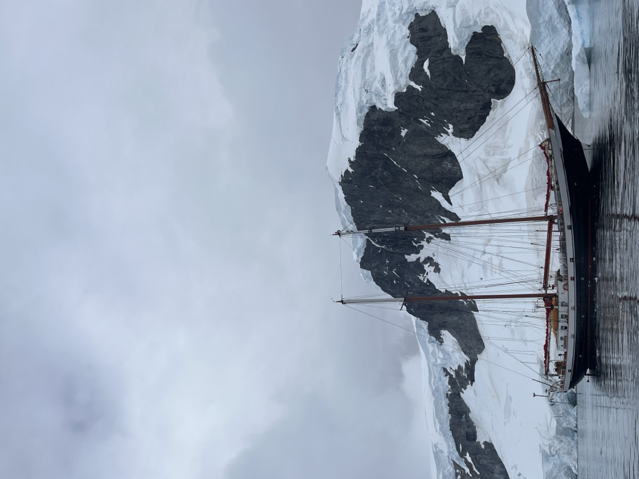 The image captures a majestic sailing vessel, likely a schooner or barkentine, positioned alongside a colossal, snow-covered mountain or glacier. The ship, with its dark hull and lighter superstructure featuring visible portholes, is equipped with two prominent masts adorned with intricate rigging and reddish-brown furled sails.
The towering backdrop consists of a rugged mountain or ice-covered landmass. Its dark, exposed rock formations are sharply contrasted by vast blankets of pristine white snow and ice that cling to its contours and extend towards the water. The immense scale of this natural formation dwarfs the ship, emphasizing the grandeur of the environment.
The ship floats on calm, dark water that subtly reflects the overcast sky and the vessel's form. The sky itself is a uniform expanse of soft grey clouds, indicating an overcast or foggy day, which bathes the entire scene in diffused, muted light. This cold, cloudy weather, combined with the immense snow and ice, strongly suggests a polar setting, such as Antarctica or the Arctic.
The activity appears to be one of quiet exploration or observation, as the ship seems stationary, perhaps anchored. There are no individuals visible on board or in the immediate vicinity, reinforcing a sense of solitude and the raw, untamed beauty of this remote landscape. The overall atmosphere is one of stark, serene beauty and adventurous tranquility. No visible text is present in the image.