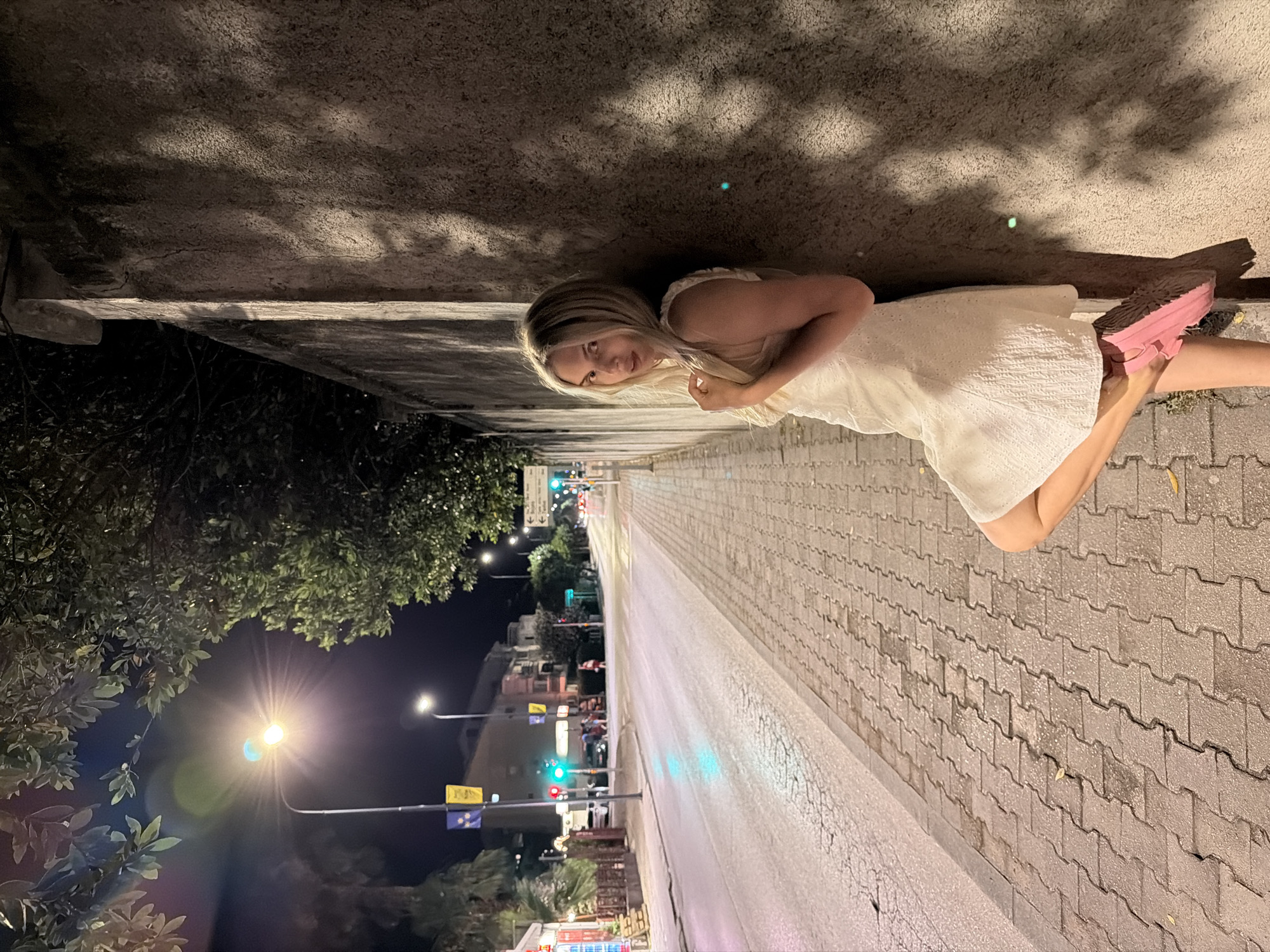 A young Caucasian woman with blonde hair, wearing a white textured dress and pink platform sneakers, leans against a textured concrete wall on a sidewalk. She is positioned on the right side of the frame, with her body angled towards the camera. Her left leg is bent and extended, with her left foot resting on the wall. She is looking towards the camera with a slight smile. Behind her, to the left, is a street scene at night. The street is paved and has a sidewalk with interlocking pavers. Streetlights illuminate the scene, casting shadows from trees onto the wall. Traffic lights are visible, with one showing red and another green. Buildings line the street in the background, and there are signs and streetlights. The overall impression is a nighttime street scene in a town or city. The scene appears to be Budva, Montenegro, given the presence of palm trees and the architectural style of the buildings, which are common in coastal Montenegrin towns.