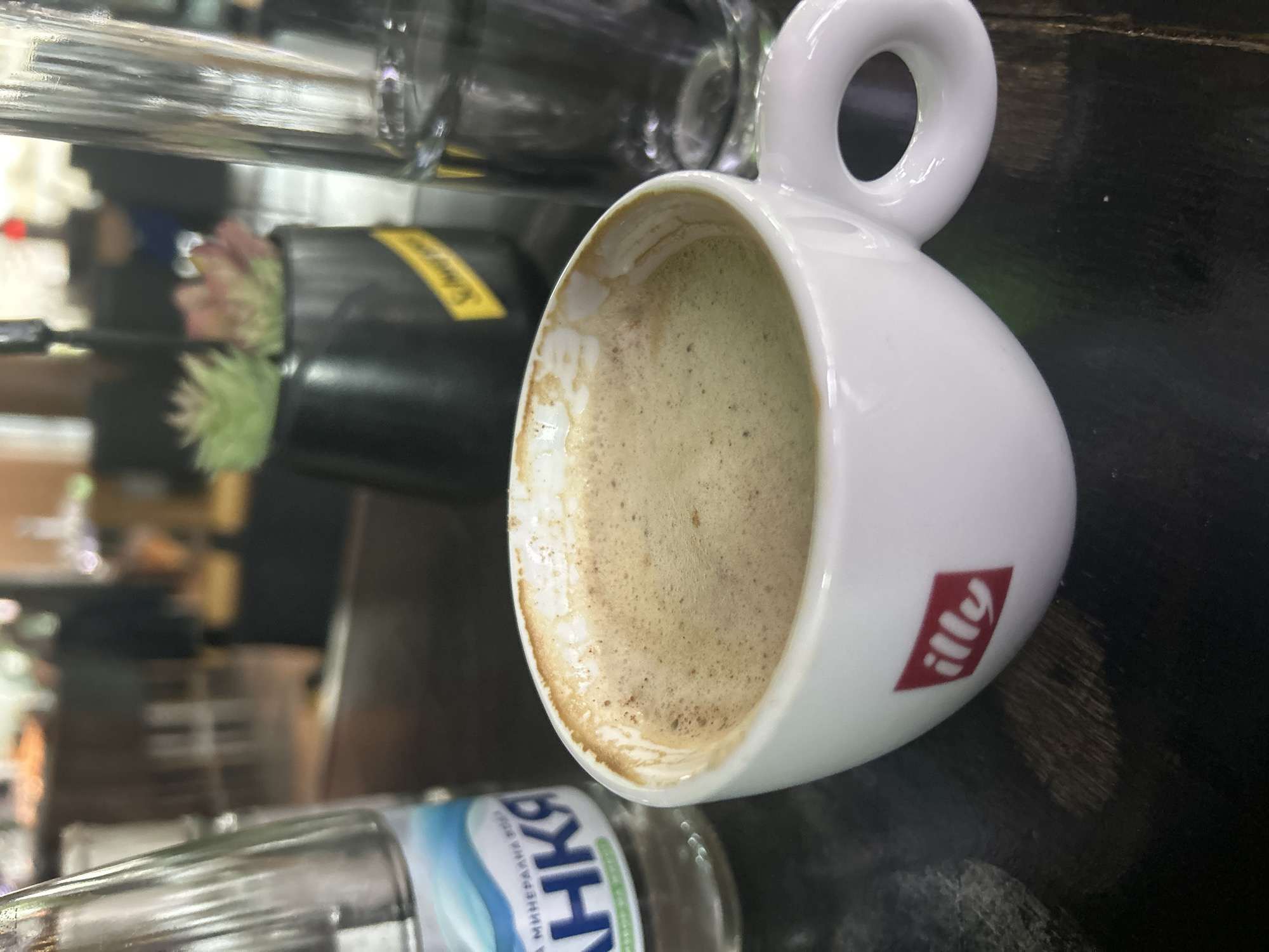 The image shows a white Illy coffee cup partially filled with coffee. The cup is on a dark surface, likely a table. A bottle of Devin mineral water is visible in the foreground. In the background, there are blurred objects including glasses, a black container with a yellow label (possibly Schweppes), and a succulent plant. The location is likely a cafe or bar in Sofia, Bulgaria based on the Cyrillic writing on the water bottle.