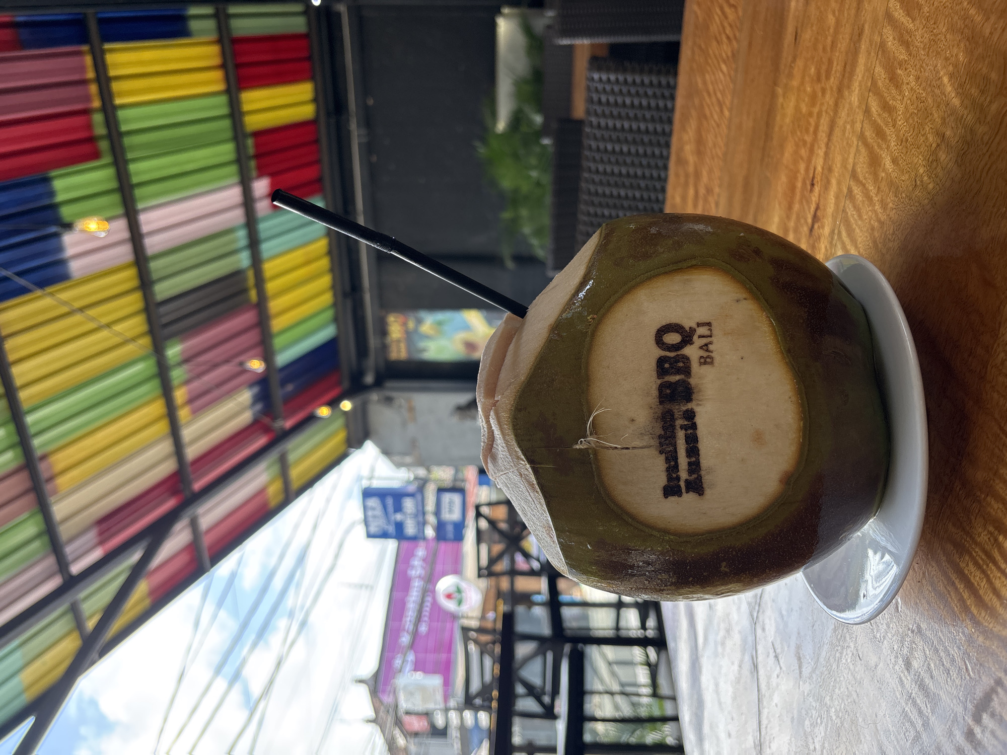 The image shows a green coconut on a white saucer with a black straw, sitting on a wooden table. The coconut is branded with the words "Brazilian Aussie BBQ BALI." The background is a blurred outdoor scene, including a building with a brightly colored, striped awning and various signs and structures. The location is likely Denpasar, Indonesia, based on the "BALI" in the coconut branding. No people are visible in the image.
