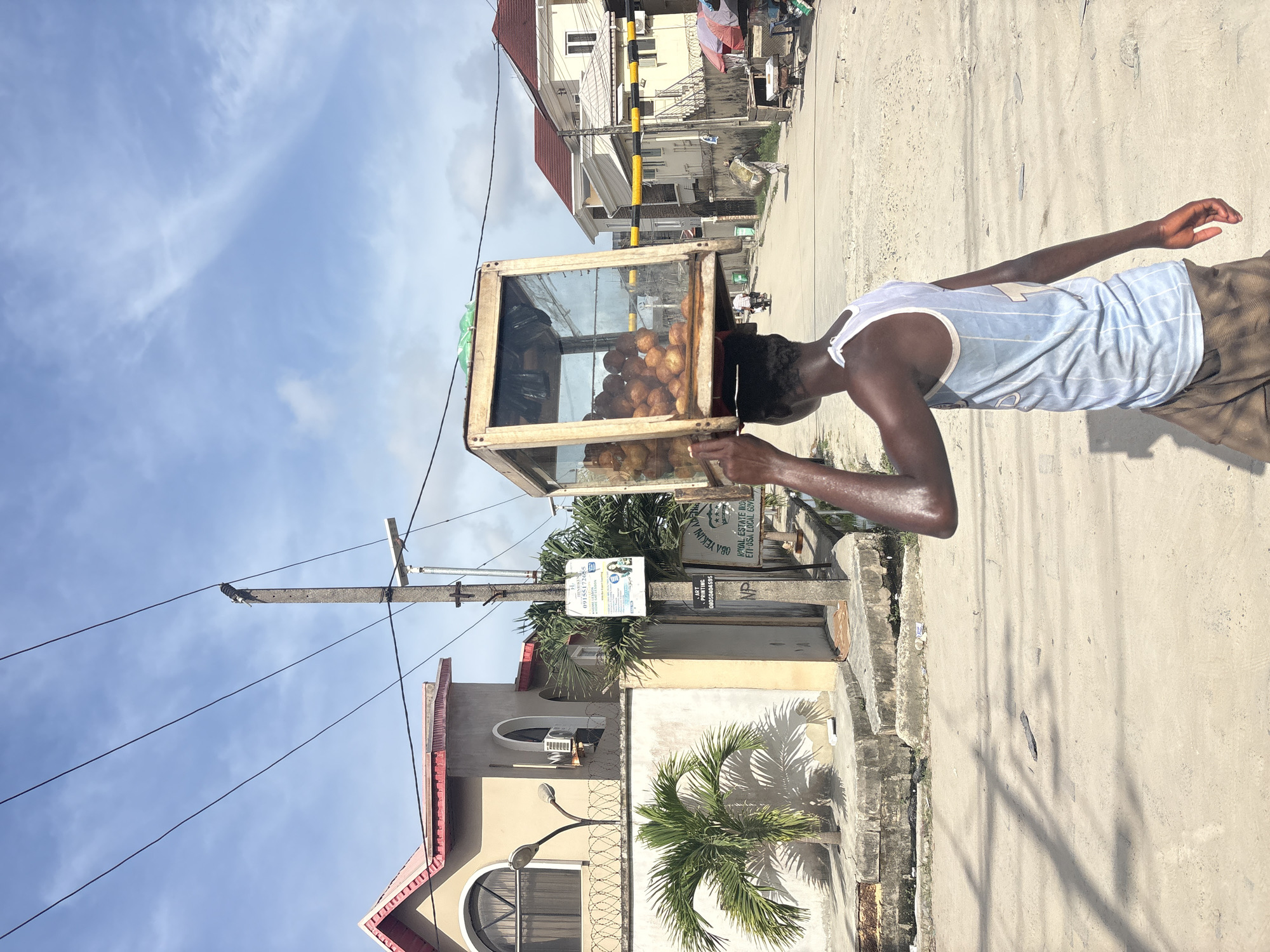 The image shows a man in Igboefon, Nigeria, carrying a glass-enclosed display case of round, golden-brown food items on his head. He is wearing a light blue and white striped tank top and brown shorts. The setting appears to be a street in Igboefon with buildings, utility poles with wires, and palm trees visible in the background. A sign on one of the utility poles has text indicating "HENRIKEN", a phone number "09155172655", the word "ART PRINTING" with a phone number "08050404595", "NP", "YEKINI ADENO", "ROYAL ESTATE IRO", and "ETI-OSA LOCAL GOVE". The sky is partially cloudy.
