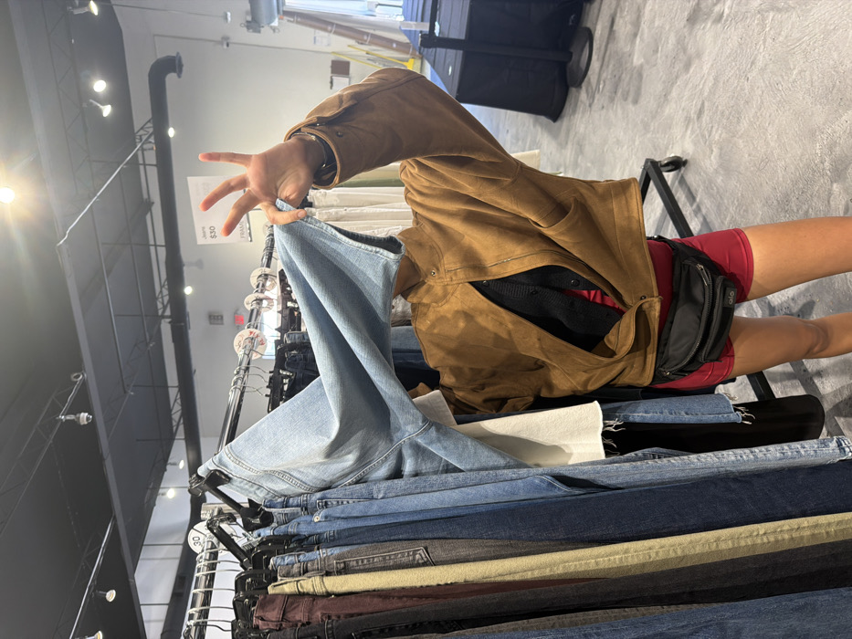 The image, originally rotated 90 degrees clockwise, captures a scene inside what appears to be a retail clothing store. The main subject is a person whose right arm, hand, torso, and legs are visible, engaged in shopping.
The person is wearing a brown, possibly suede or leather, jacket with visible snap closures. Underneath the open jacket, a black garment (possibly a sweater or cardigan) and a red garment are layered. The lower body shows red shorts or biker shorts, and a black fanny pack or waist bag is worn around the waist/hip, positioned towards the back right side. The person's right hand is prominently displayed in the upper part of the image, holding up a pair of light blue denim jeans from a clothing rack. The hand is open with fingers slightly splayed, as if examining the fabric or fit of the jeans.
The foreground and midground are dominated by multiple clothing racks filled with various pairs of jeans. These jeans come in different washes and colors, including light blue, medium blue, dark blue, black, and possibly some earthy tones, all neatly folded or hung. The metal rails and hangers of the clothing racks are clearly visible.
The setting is an indoor commercial space, likely a clothing store or a section of a department store. The store features a high ceiling with exposed industrial elements such as pipes, beams, and track lighting fixtures, some of which are illuminated and appear as bright light sources. The floor is a light gray, concrete-like material. In the background, dark, box-like structures (possibly speakers or other equipment) can be seen.
Visible text includes "S30" and "FRAME" on a white tag attached to one of the clothing racks, with "$30" also discernable nearby. The overall activity depicted is a person browsing or selecting clothing in a store environment.