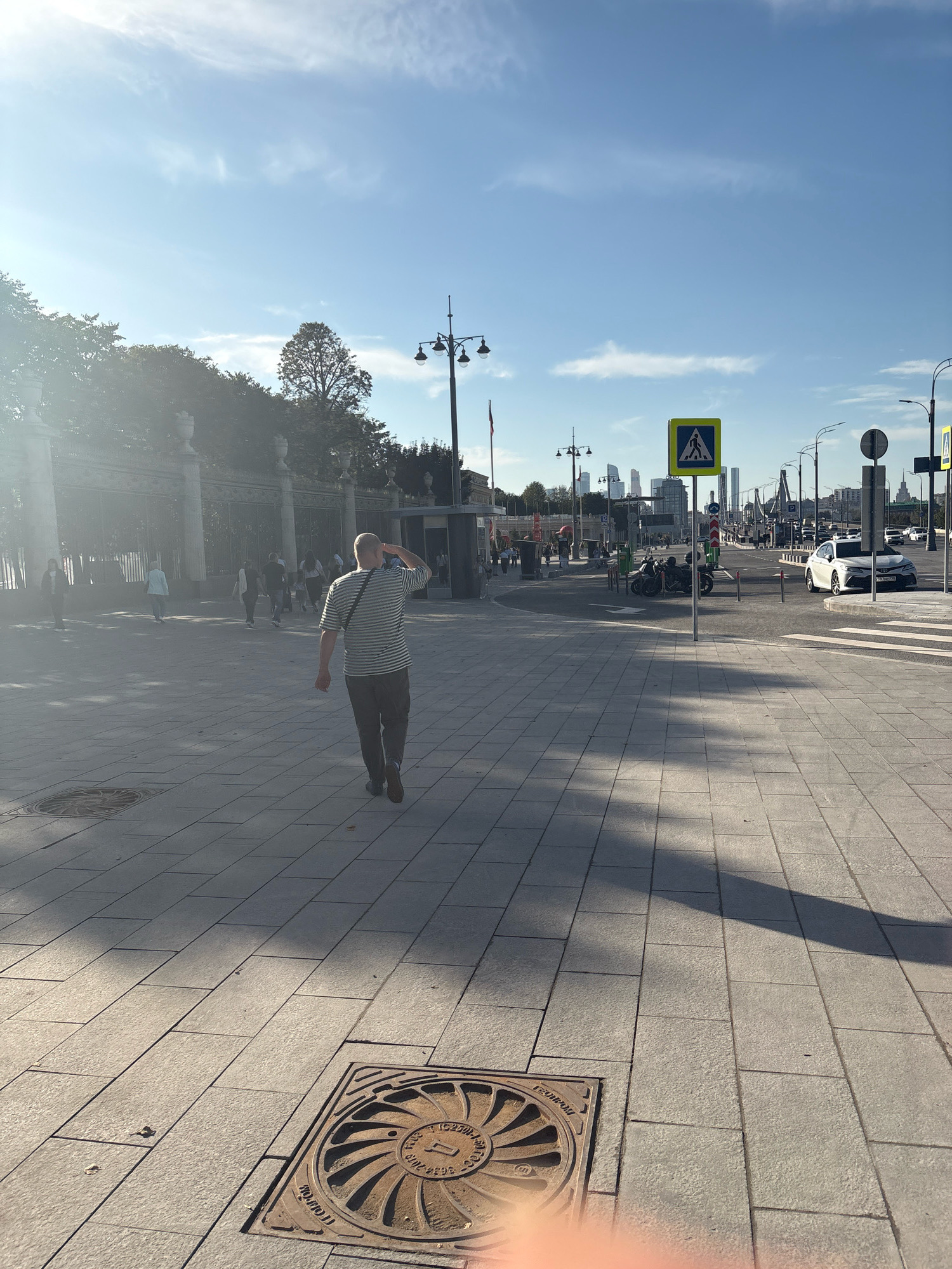 A man in a striped shirt and dark pants is walking on a paved plaza in Moscow, Russia. He is saluting the camera or looking up. The plaza has large, square paving stones, and a manhole cover is visible in the foreground. In the background, there is a street with cars, streetlights, and pedestrian crossing signs. Several other people are walking in the distance, and a tall fence with ornate posts lines the left side of the plaza. The sky is clear and blue, with some wispy clouds. The sun is casting long shadows across the plaza. The overall impression is of a sunny day in a public urban space.
