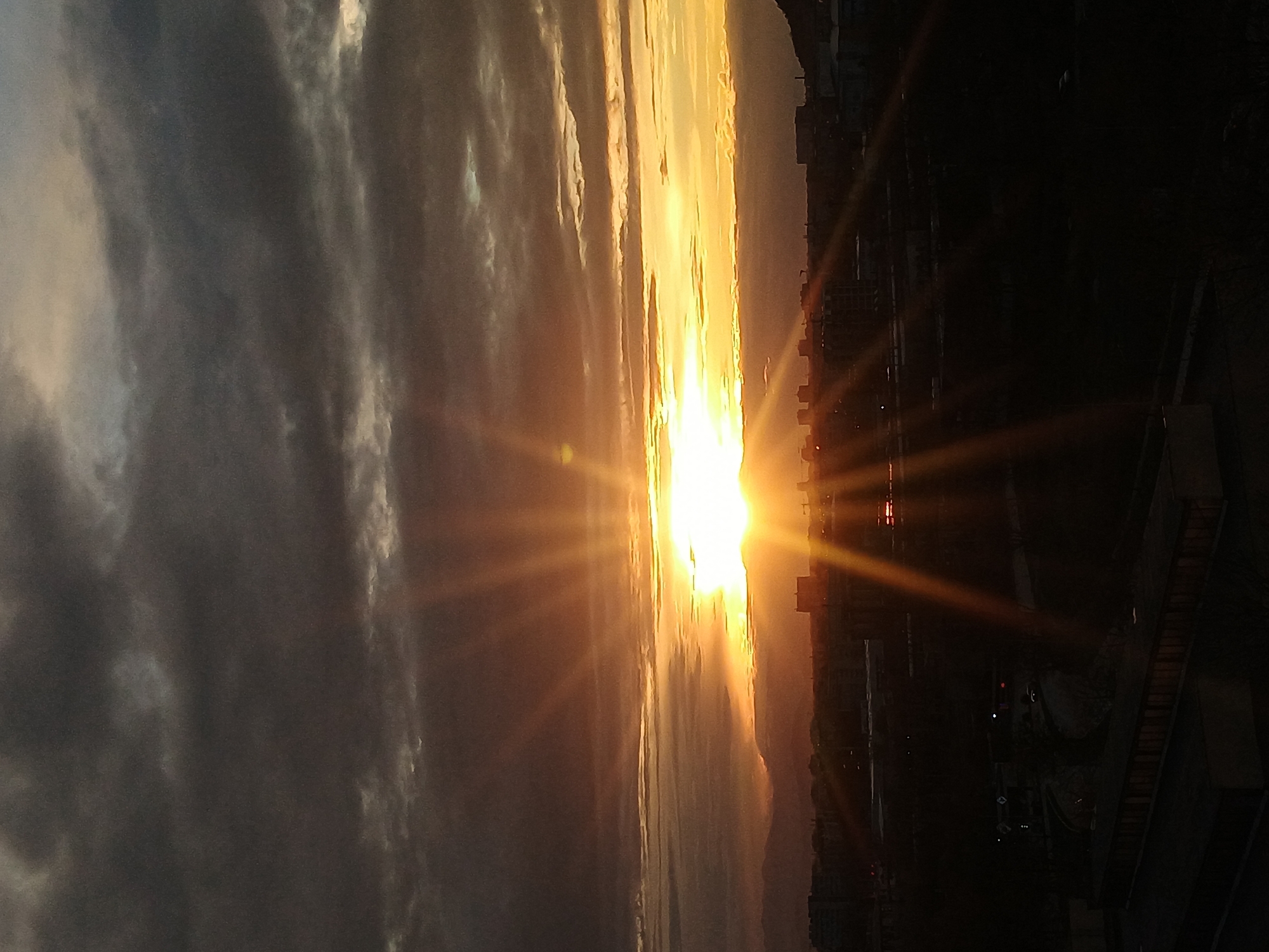 The image captures a sunset in Plovdiv, Bulgaria, with the sun positioned centrally, radiating golden light. A city skyline is silhouetted against the bright sky, showing buildings and urban infrastructure. A cloud formation on the left contrasts with the sunlit areas. The image has a rotated orientation.