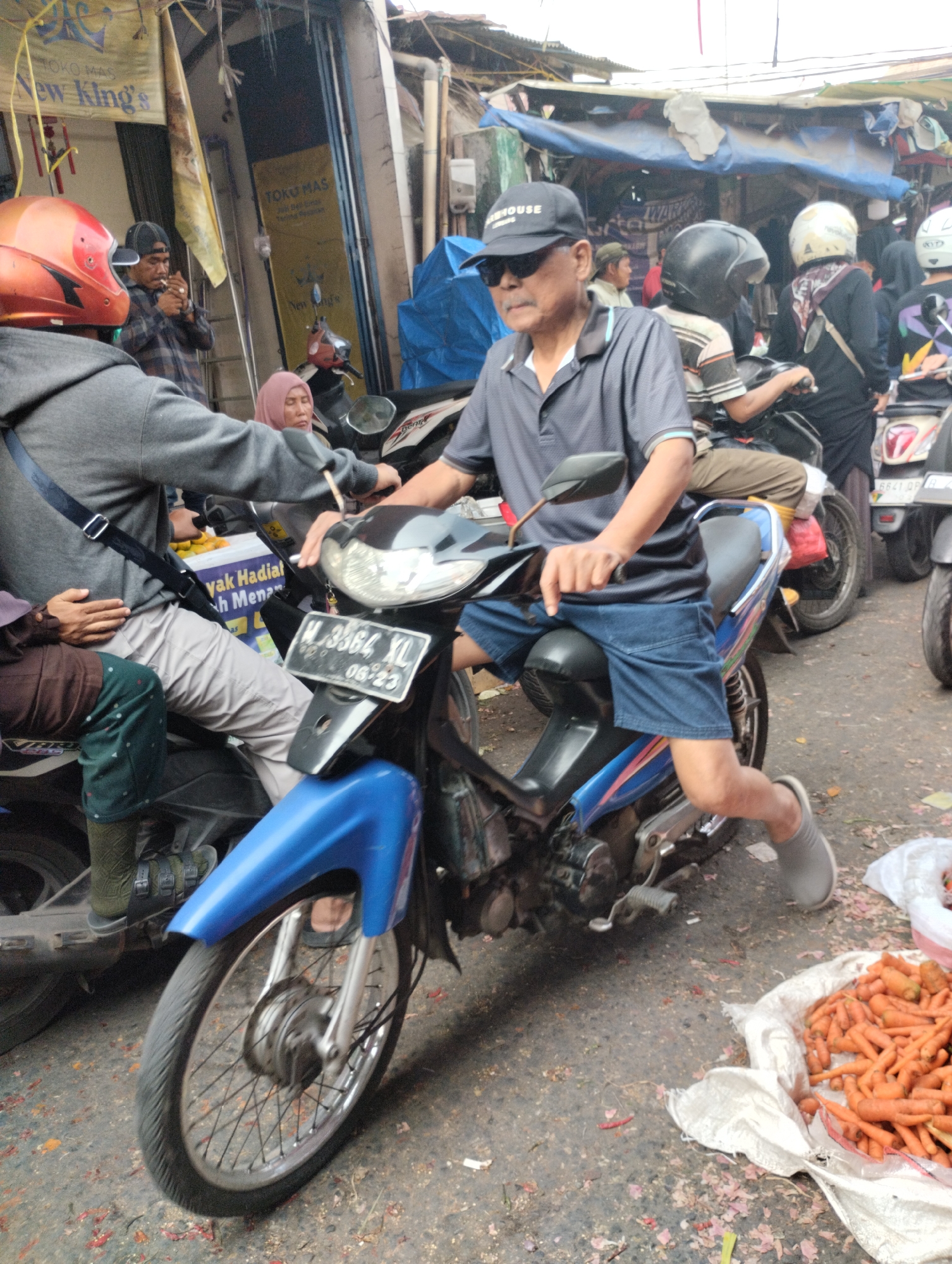 A middle-aged Asian man wearing a dark grey polo shirt, blue denim shorts, sunglasses, and a black baseball cap is seated on a blue and silver motorcycle. He is holding the handlebars with his right hand and appears to be in motion or about to move. The motorcycle has a license plate reading "M 3364 XL 08-23". In the background, there are other individuals on motorcycles, market stalls with yellow signs that read "TOKO MAS" (Gold Shop), and a pile of carrots in a white bag on the ground. The scene appears to be a busy street market in Bekasi, Indonesia, judging by the signage and the overall atmosphere of the environment. The ground is paved and littered with debris.
