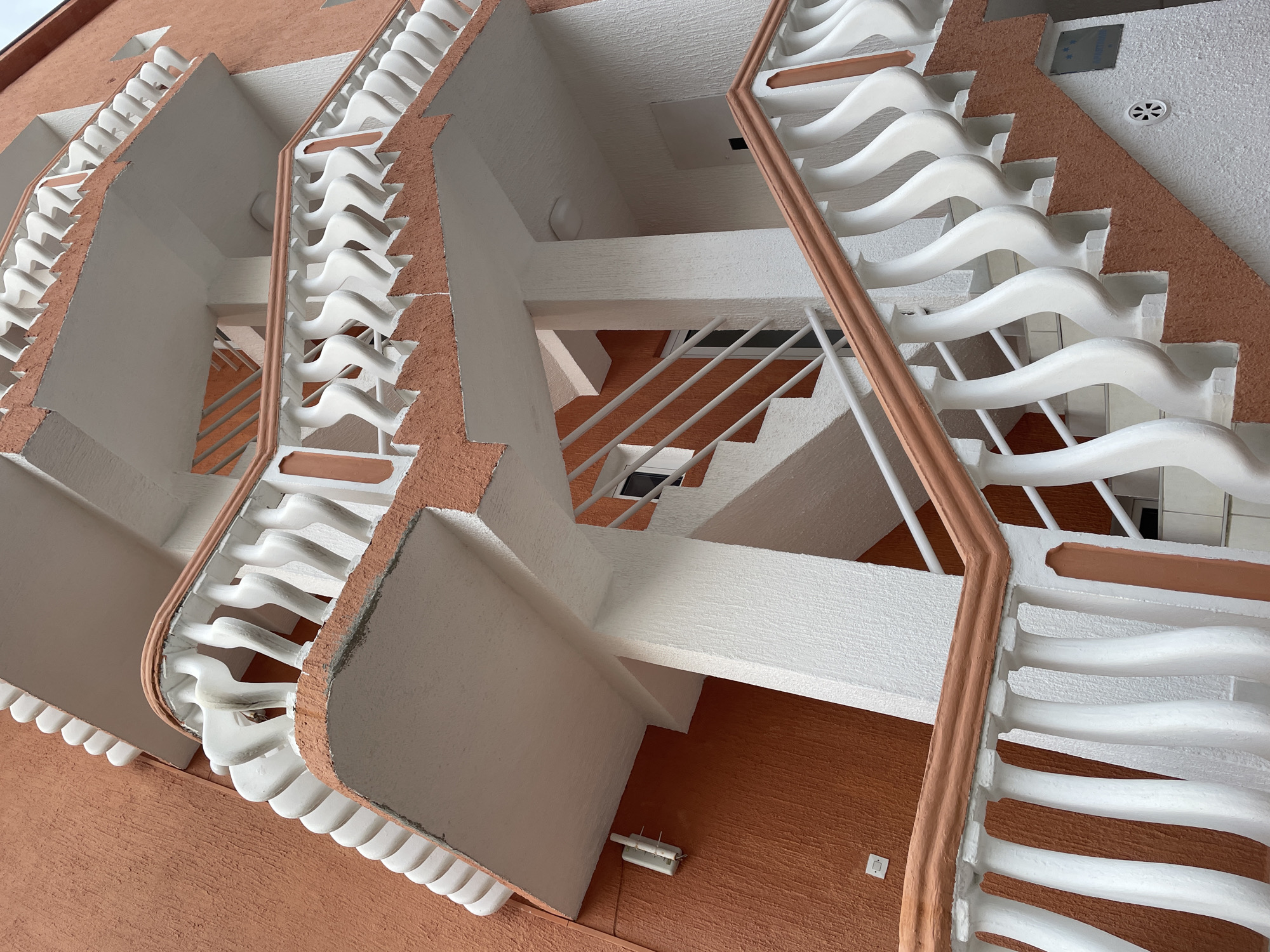 The image depicts an upward-looking view of the exterior staircases of a building in Rafailovići, Montenegro. The building has an orange textured exterior, with white decorative elements. The staircases feature white, undulating balusters and reddish-brown handrails. The staircases are interconnected, creating a complex, geometric pattern against the sky. The structure's architecture is modern and appears to be part of a residential or hotel complex. No people are visible in the frame.